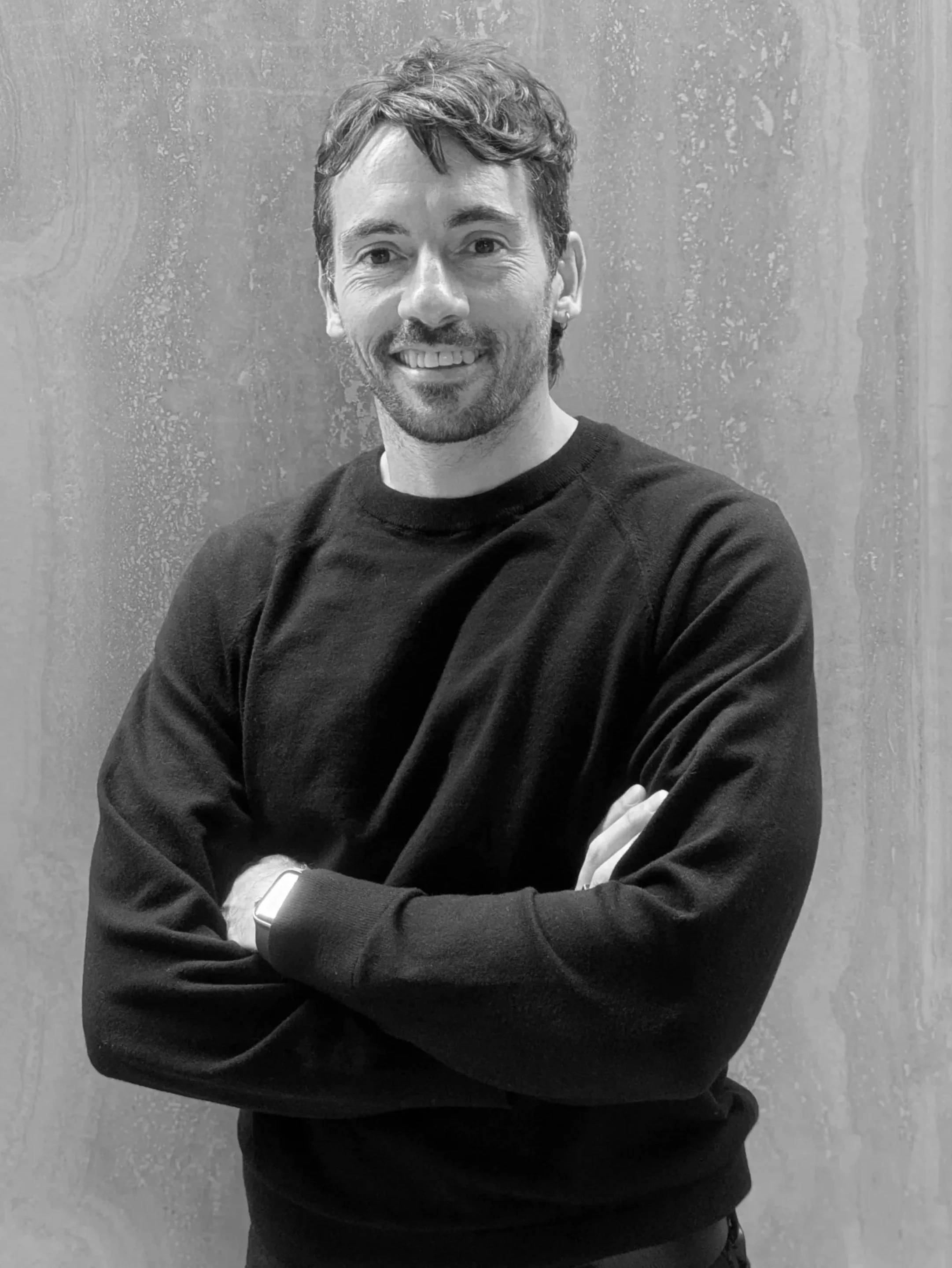 A smiling man with light skin, wavy dark hair, and a beard, wearing a black sweater, with arms crossed, standing in front of a textured wall.