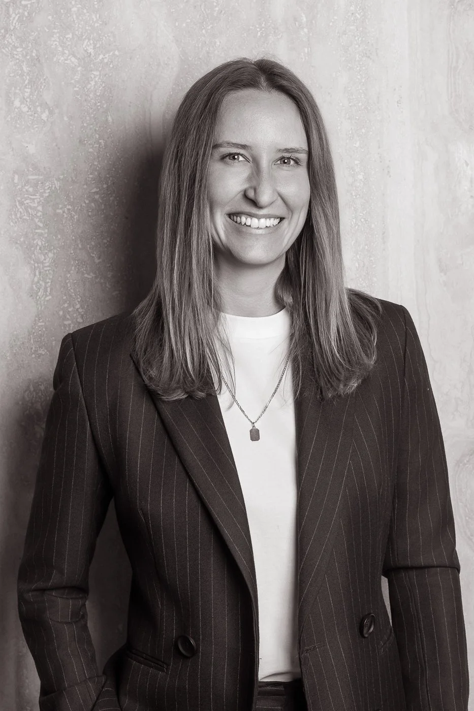 Black and white portrait of a woman with shoulder-length hair, smiling, wearing a pinstripe blazer, a white shirt, and a necklace, standing against a textured wall.