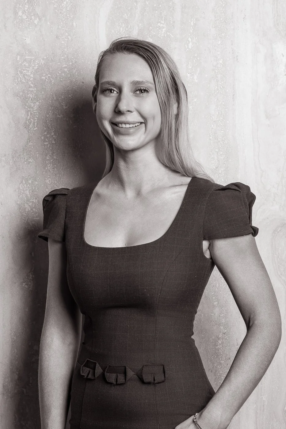 A black-and-white portrait of a smiling woman with long hair, wearing a fitted dress with puffed shoulder sleeves and decorative bows at the waist, standing against a textured wall.
