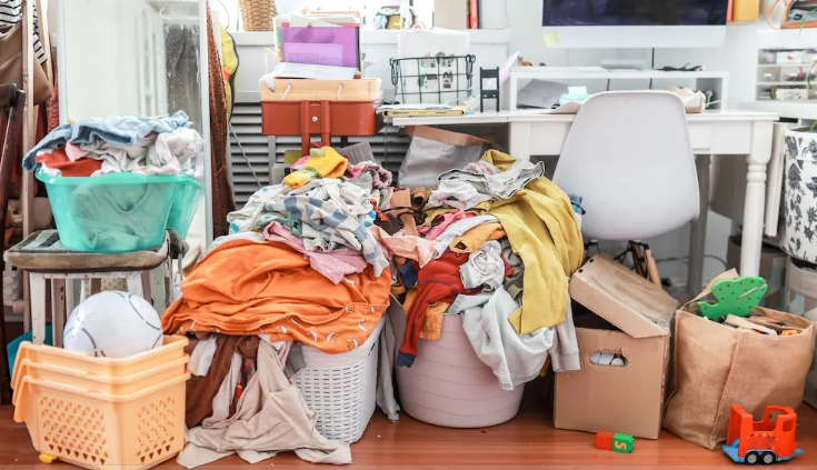 A pile of assorted laundry and clothing cluttered on the floor of a home office or craft room, with various storage containers, a chair, and craft supplies in the background.