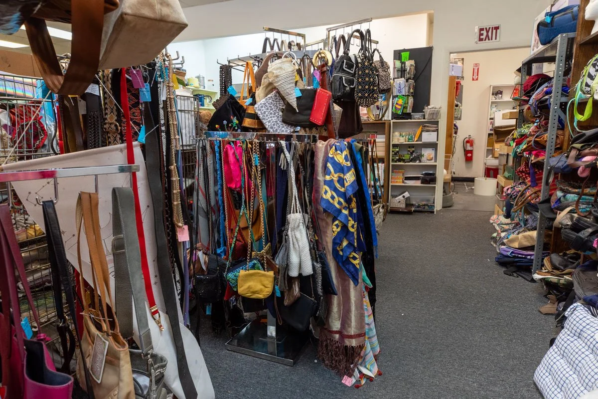 Clothing accessories and bags displayed on racks and shelves inside a thrift store, with an exit door and fire extinguisher visible in the background.