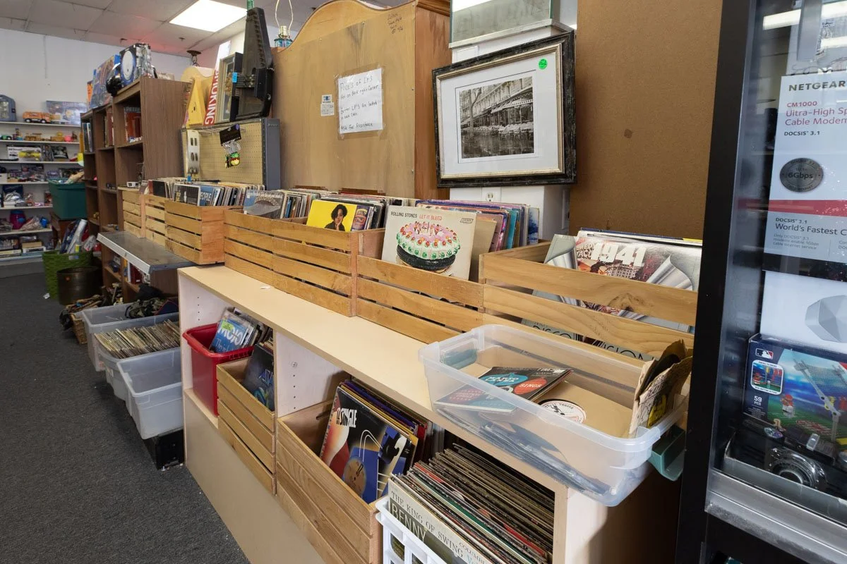 Shelves filled with vinyl records, some in wooden crates and plastic boxes, in a secondhand shop, with framed black-and-white photos on the wall and a vending machine to the right.