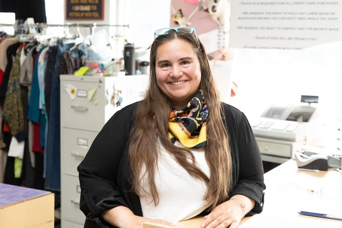 Smiling woman with long brown hair, wearing glasses on her head, a black cardigan, white top, and a colorful scarf, standing in an office or retail environment with clothing racks and office equipment in the background.