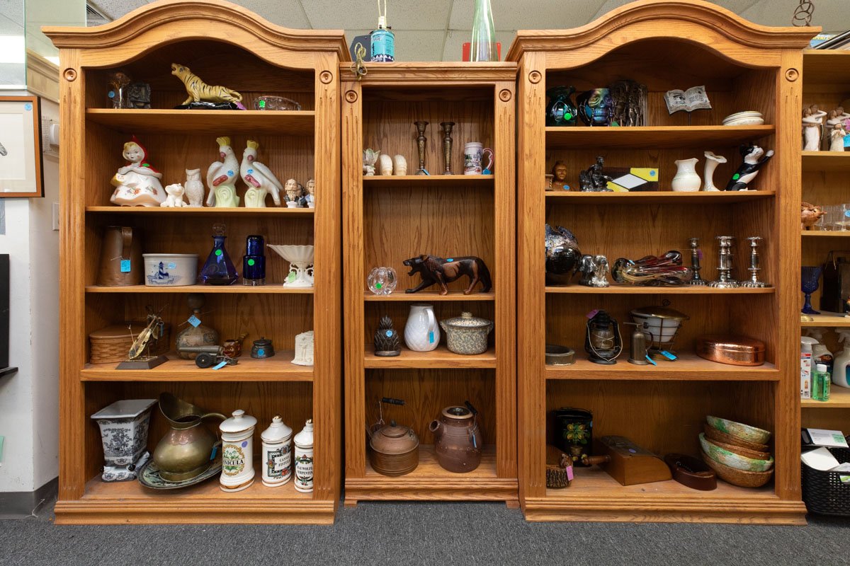 Three wooden bookshelves filled with decorative and historical items, including ceramics, glassware, figurines, vases, and sculptures, arranged on multiple shelves.
