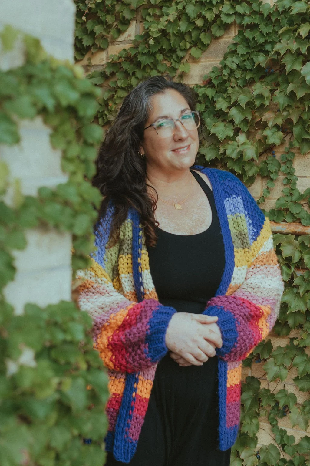 A woman with dark, wavy hair and glasses standing outdoors in front of a wall covered with green ivy, wearing a colorful knitted cardigan over a black top.
