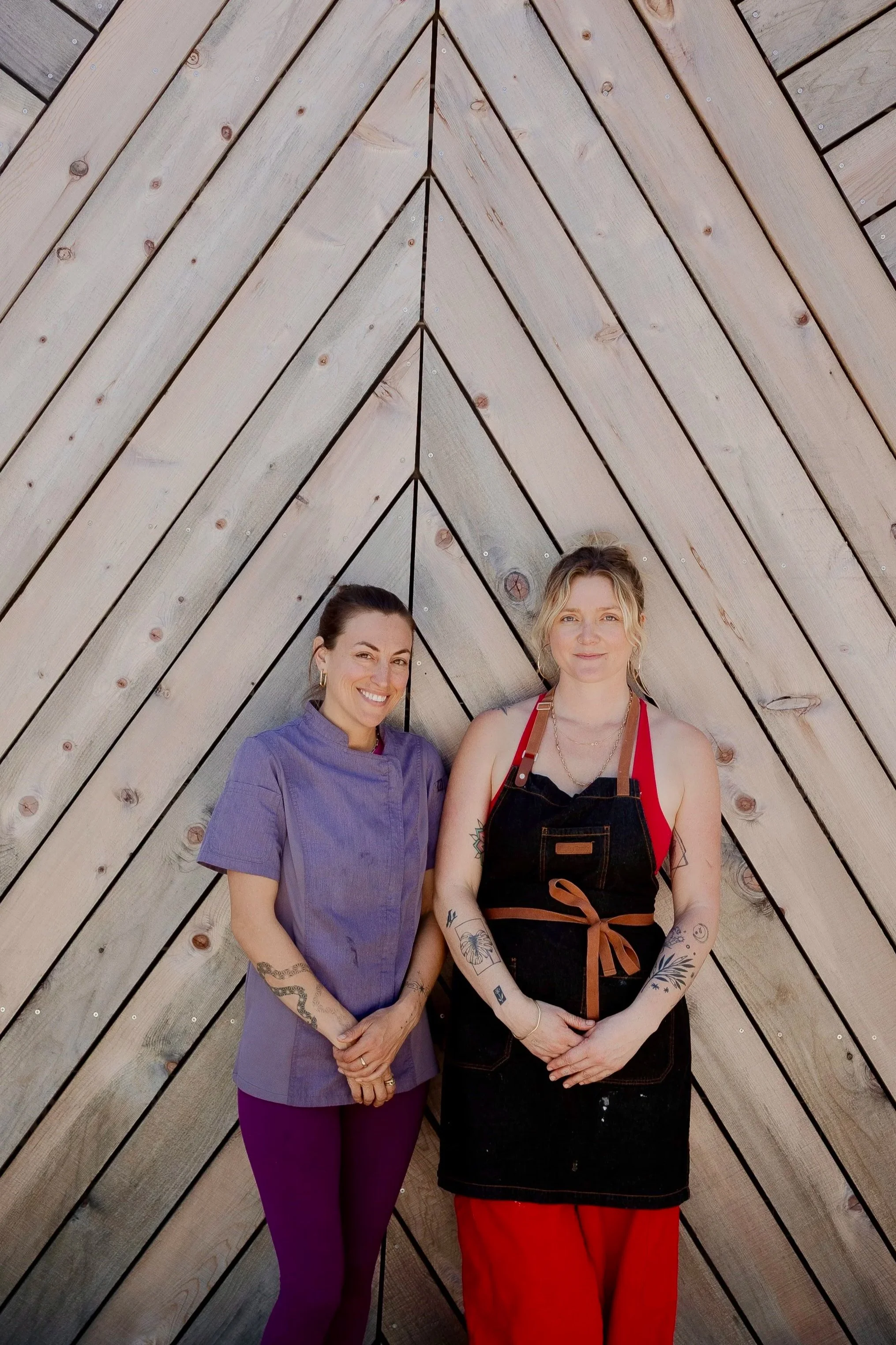 Two women standing in front of a wooden wall, smiling at the camera.