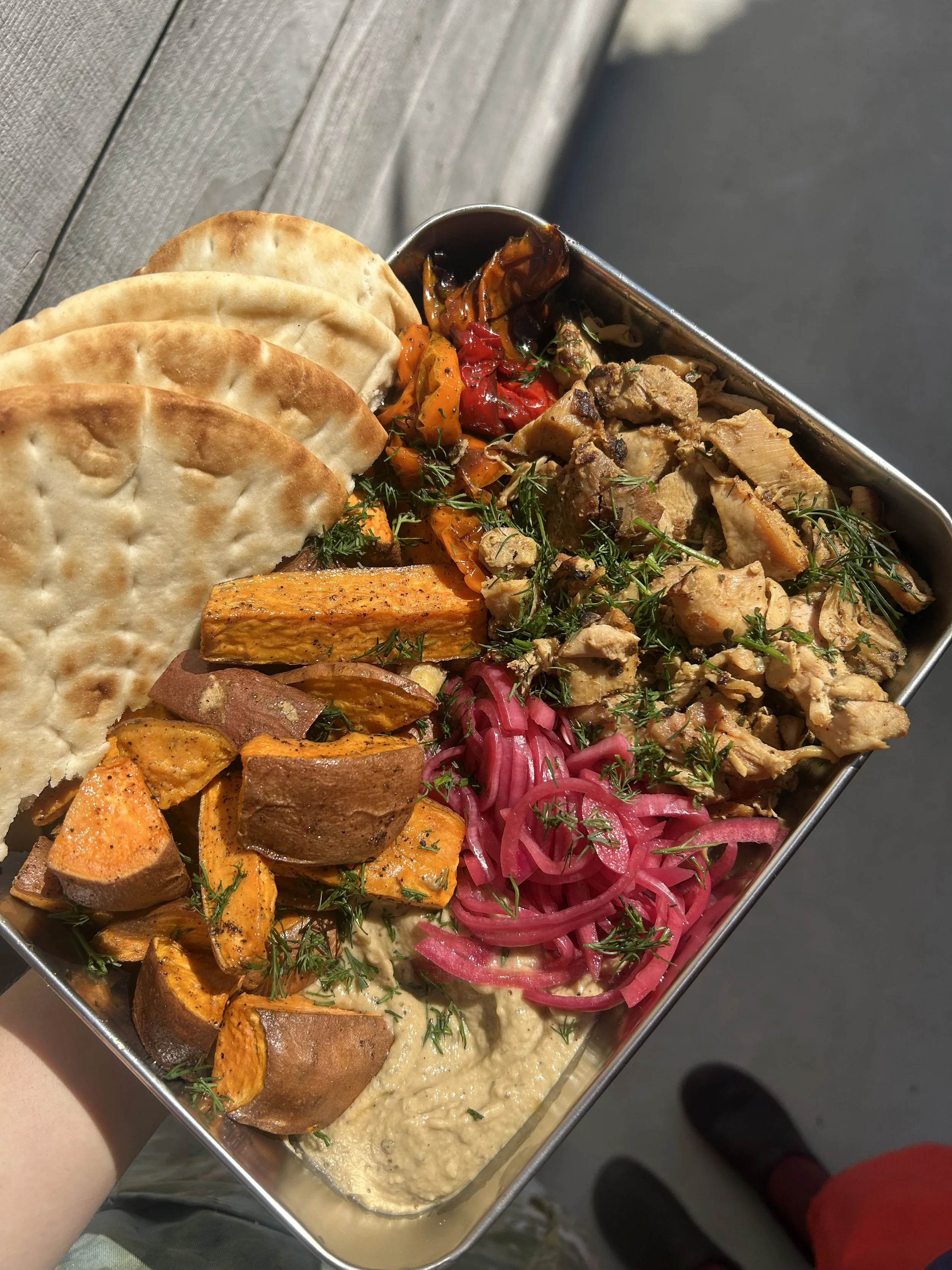 A metal tray of assorted Middle Eastern food, including pita bread, roasted sweet potatoes, pickled onions, hummus, grilled chicken, roasted vegetables, and a vegetable medley.