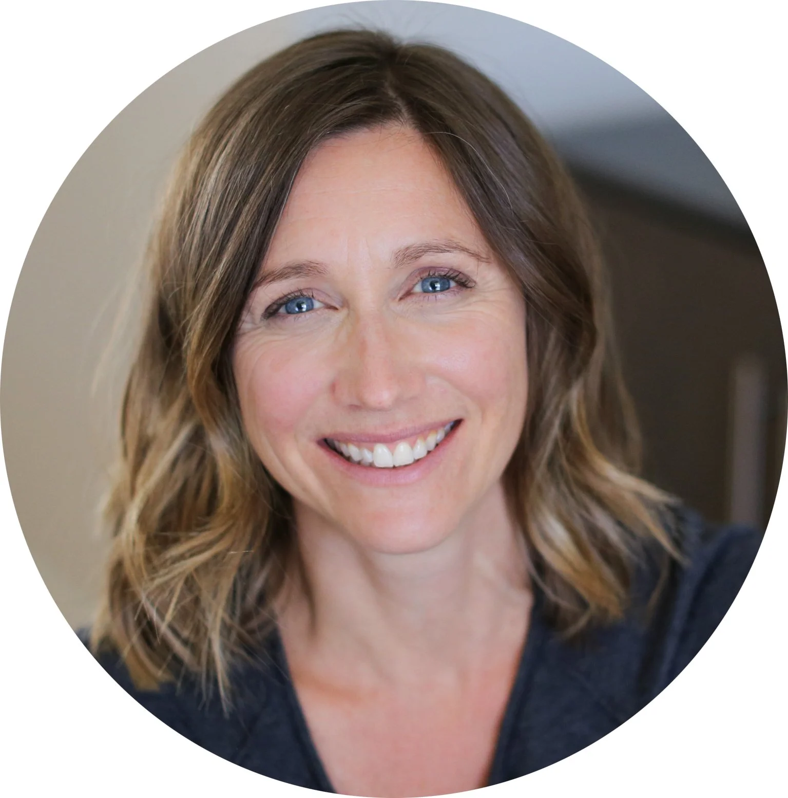 A smiling woman with shoulder-length brown hair and blue eyes, wearing a dark shirt, in an indoor setting.