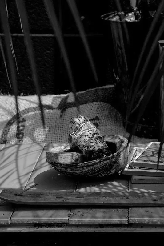 A shell on a wooden plate with a string around it, placed inside a large seashell, on a tiled surface with shadows from nearby plants.