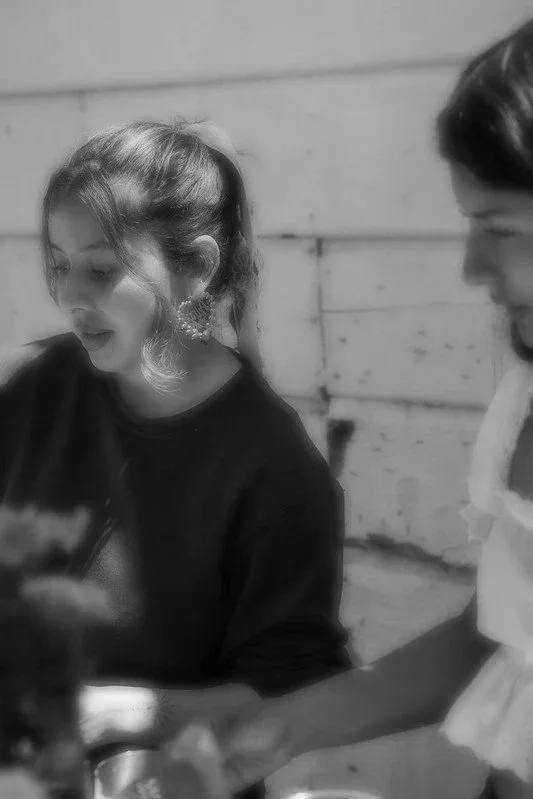 A young woman and a young girl sit together, looking at something in front of them, in a black and white photo. The woman has long hair and large earrings, and the girl has her hair tied back.