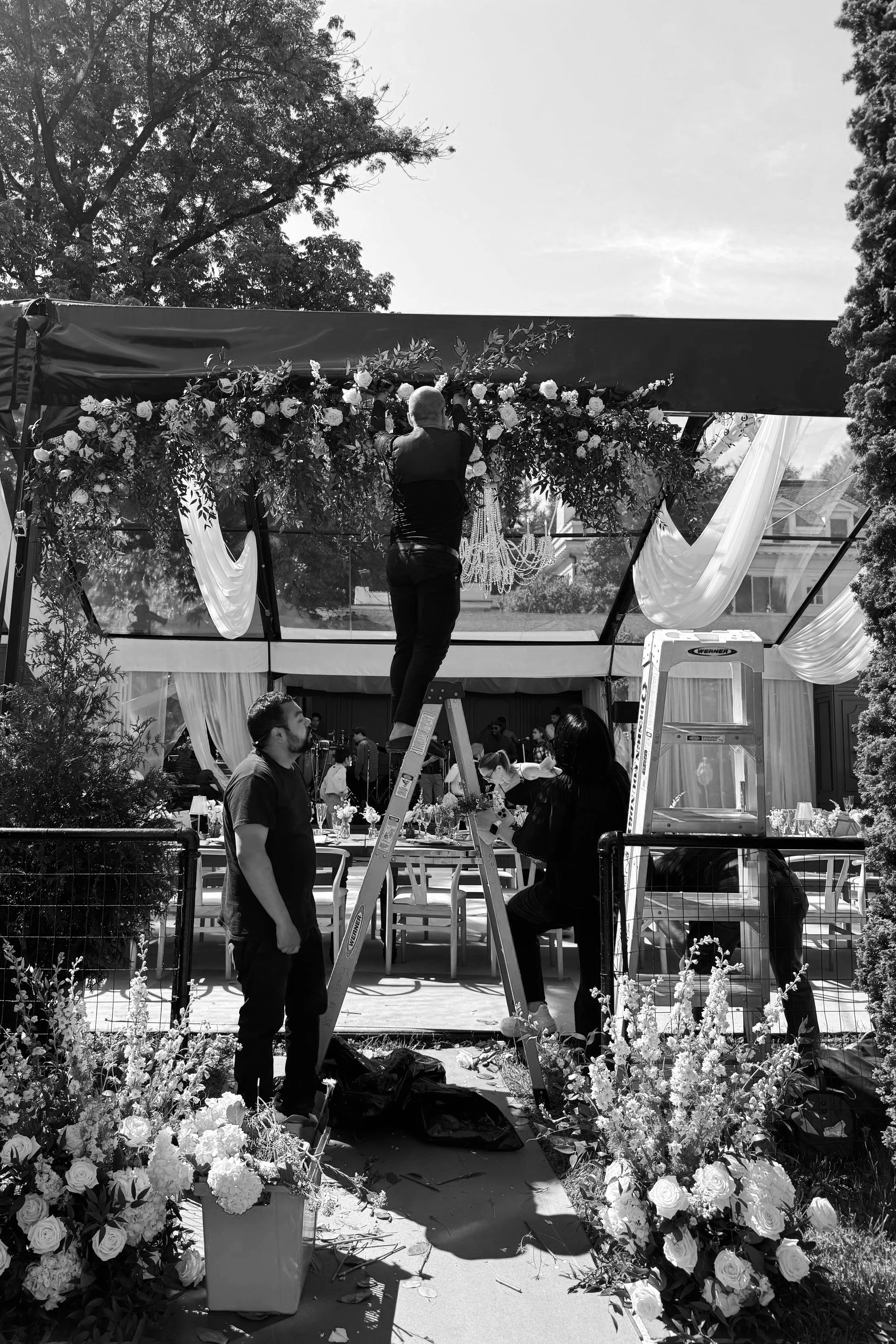 People decorating a wedding reception space with flowers and drapery, some standing on a ladder, outdoors with trees and a partly cloudy sky.