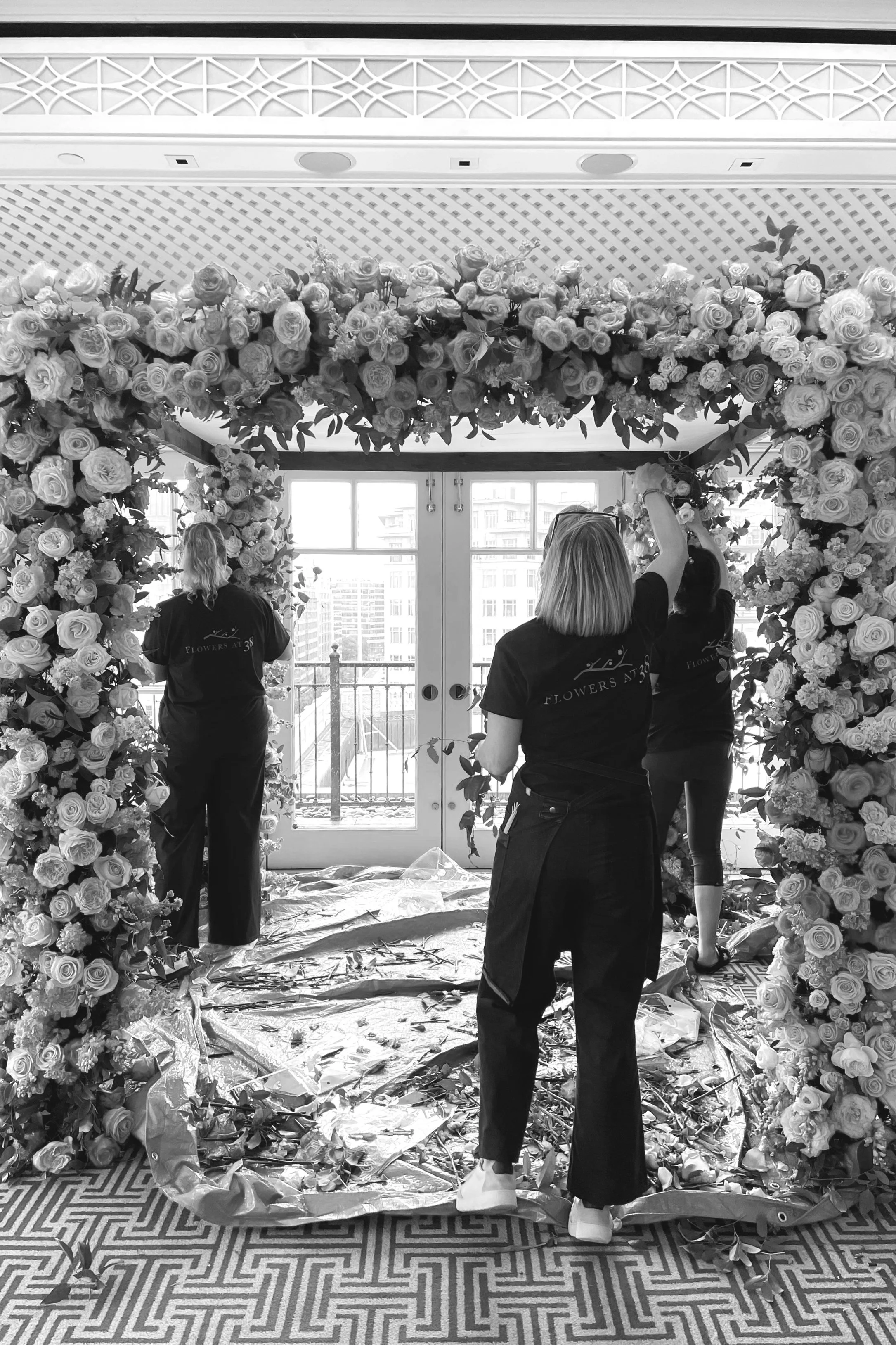 Three women decorating a floral arch with roses and foliage inside a room near large glass doors, with a cityscape view outside.