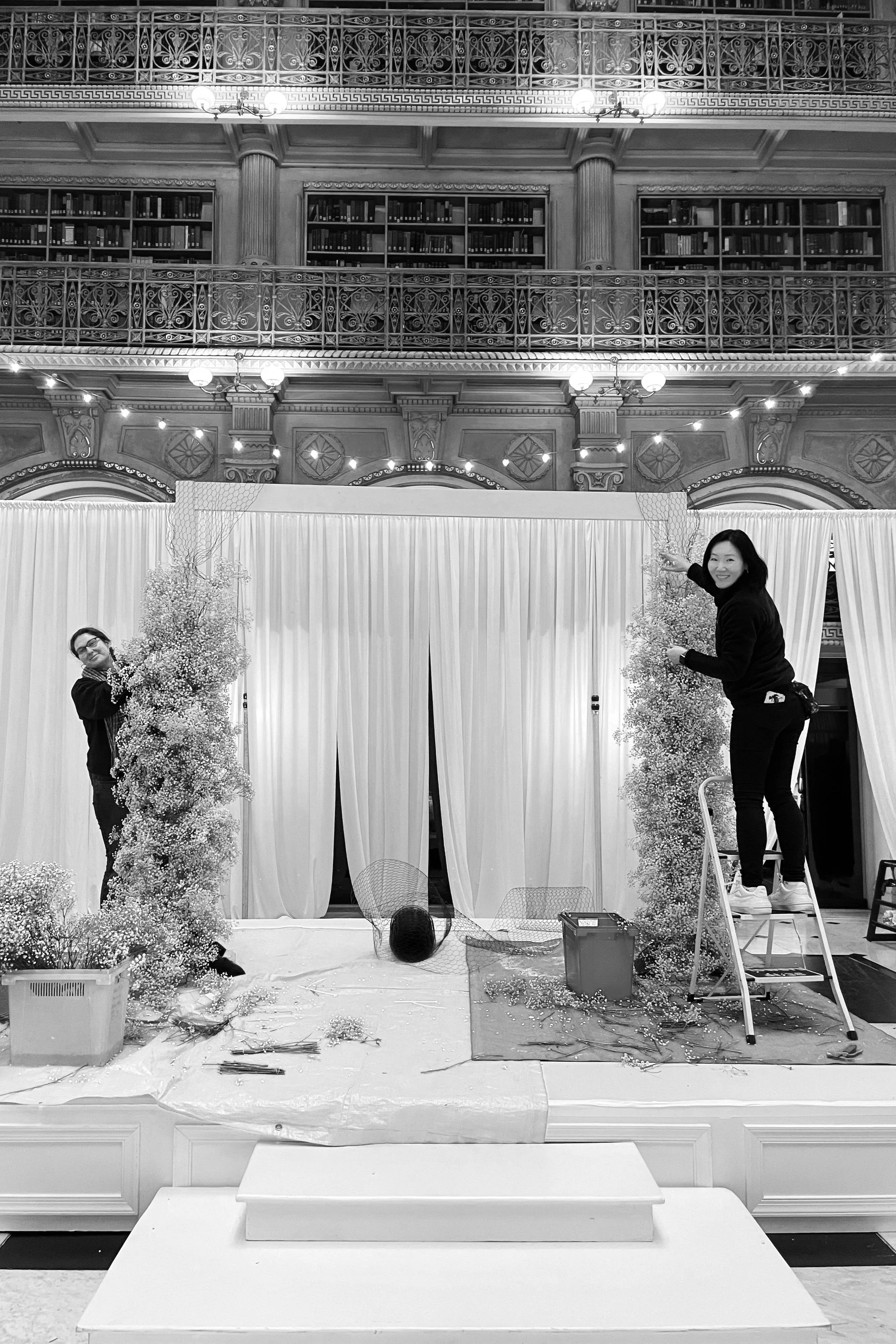 Two women decorating a stage with trees and flowers in a grand hall with ornate railings and bookshelves in the background, using a ladder and standing on the stage.