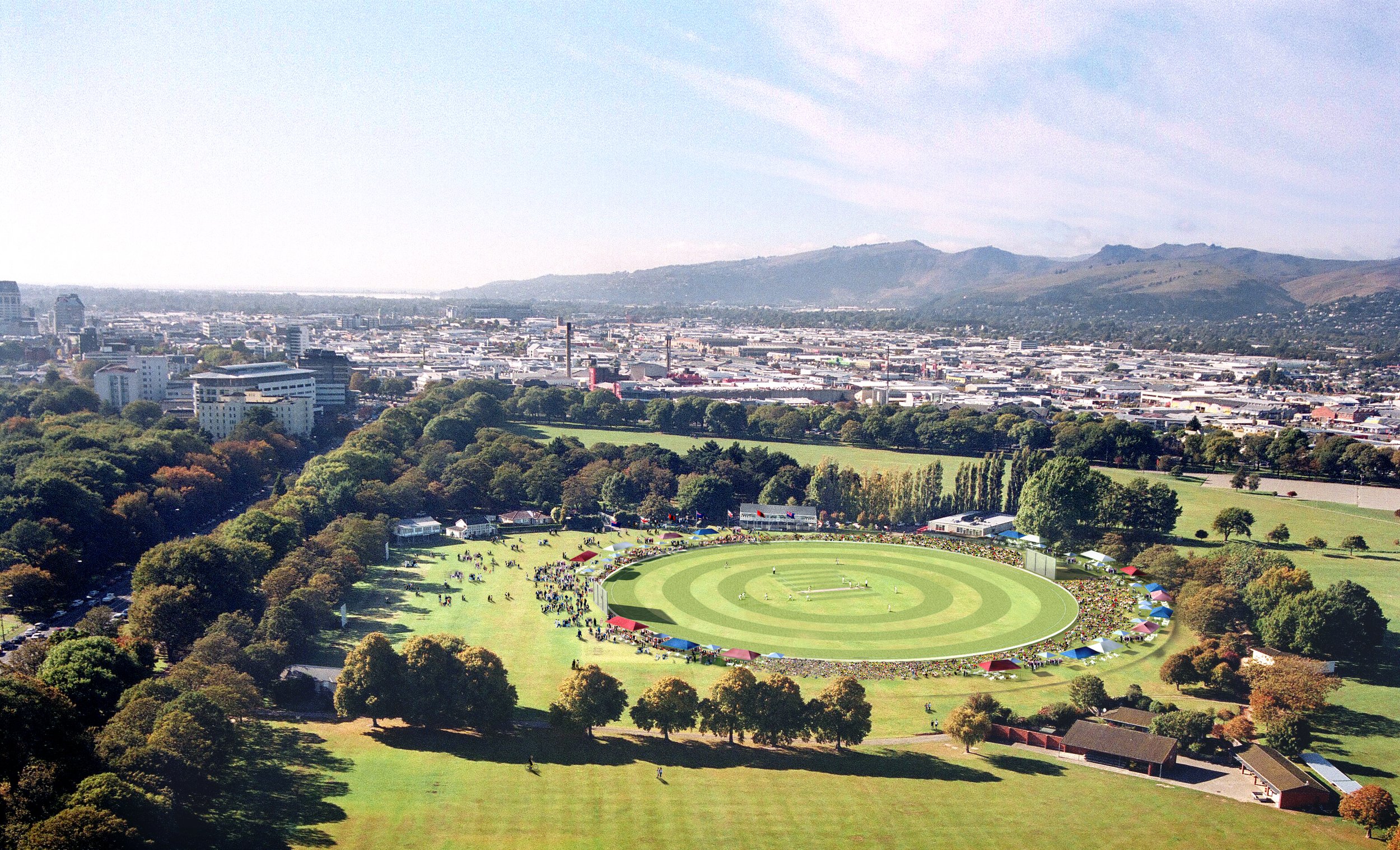 Aerial view of a cricket ground with spectators, surrounded by trees and city buildings in the background, and mountains in the distance.