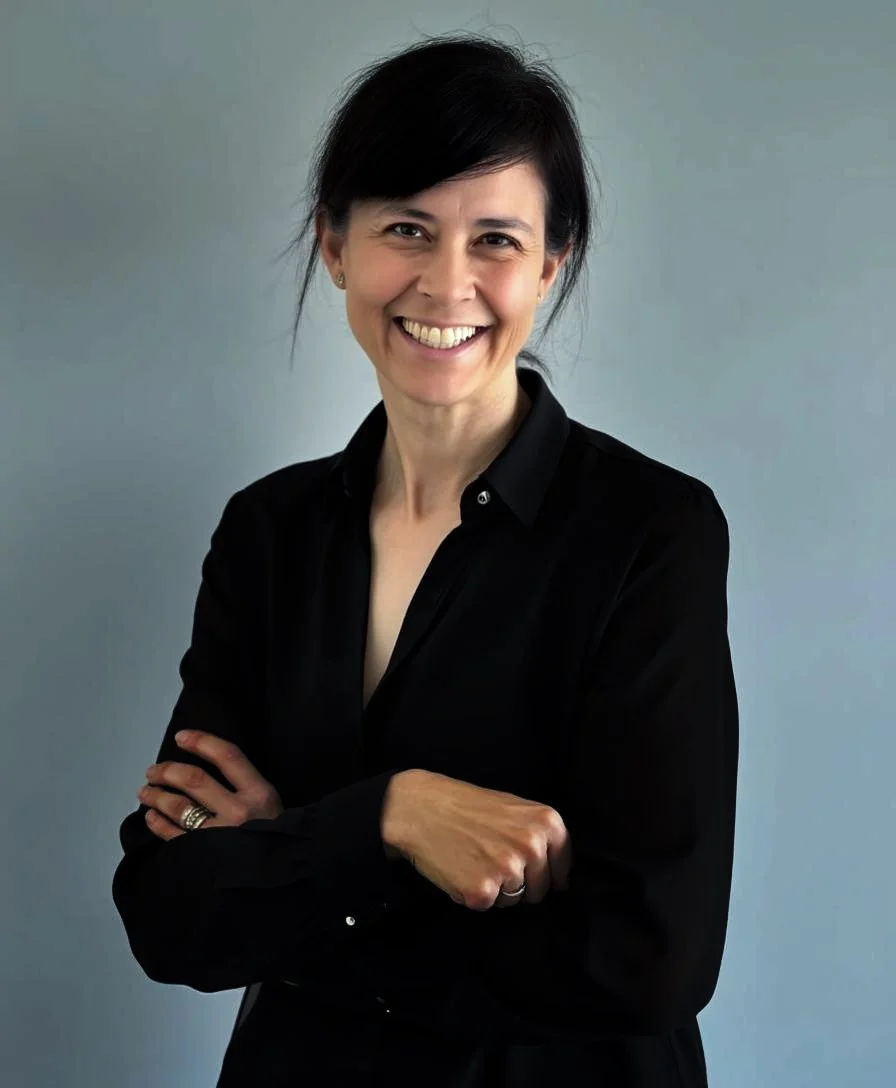 A smiling woman with black hair in a black shirt standing with arms crossed against a plain gray background.