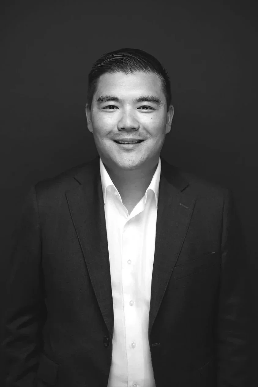 Black and white portrait of a man in a suit smiling, against a dark background.