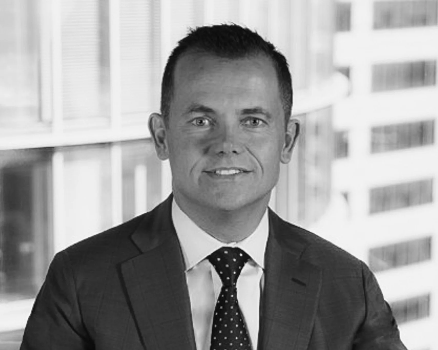 Black and white professional portrait of a man in a suit and tie, in an office setting with a large window and city buildings in the background.
