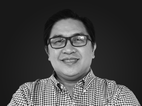 A man with dark hair, glasses, and a checkered shirt smiling at the camera against a black background.