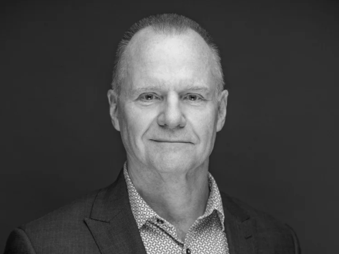 Black and white portrait of a middle-aged man with short hair, wearing a suit jacket and patterned shirt, looking directly at the camera with a slight smile, against a plain dark background.