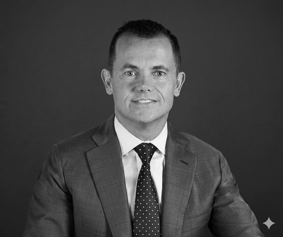 Black and white professional portrait of a man in a suit and tie, in an office setting with a large window and city buildings in the background.