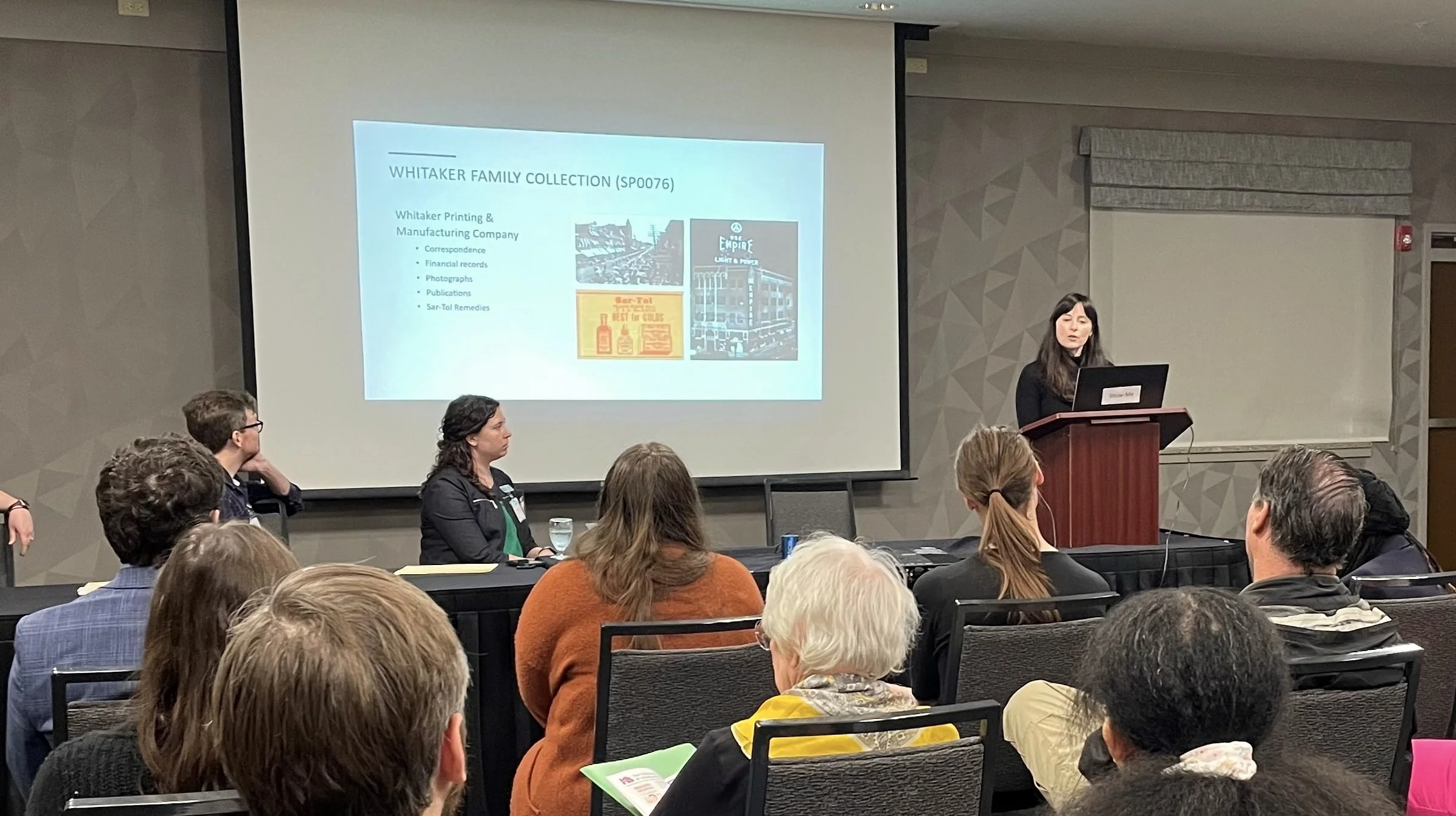 A woman presenting at a conference with a slide about the Whitaker Family Collection displayed on the screen. The room is filled with seated attendees listening attentively.