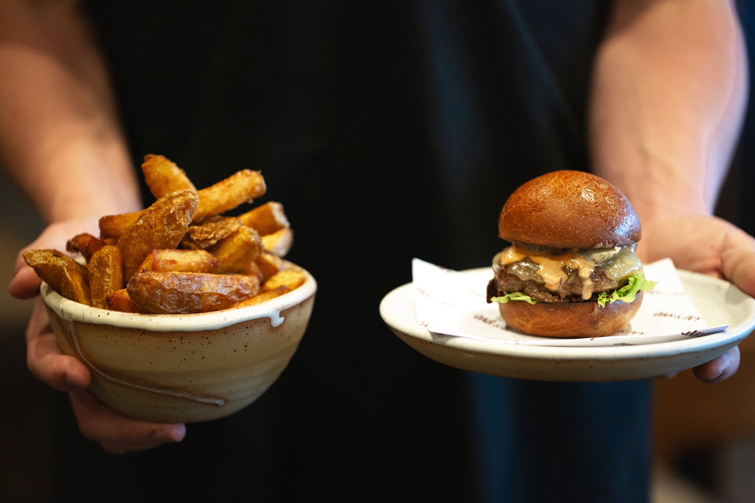Person holding a bowl of French fries in the left hand and a plate with a cheeseburger in the right hand.