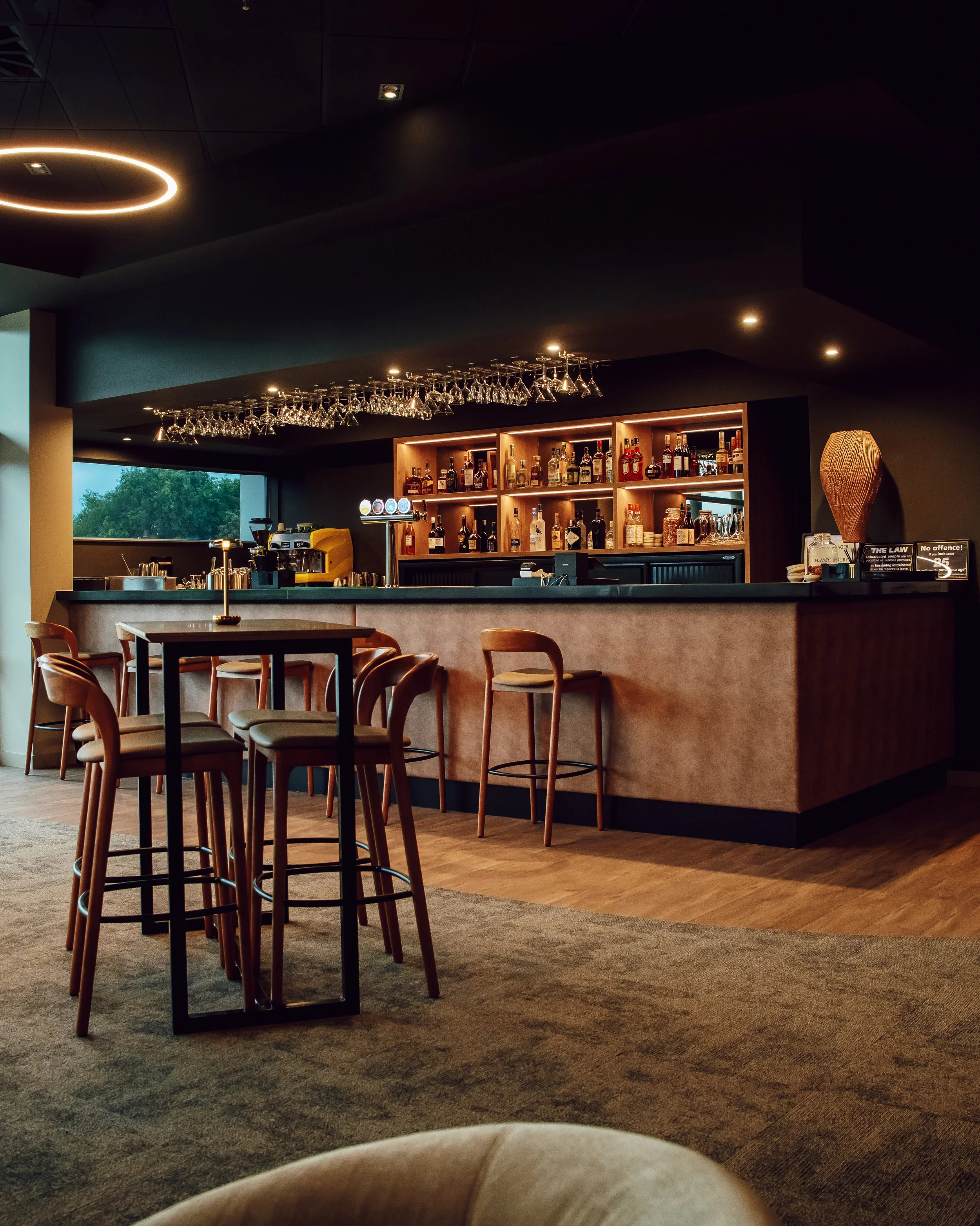 A modern bar with a black ceiling, hanging wine glasses, a wooden bar counter with a variety of liquor bottles on illuminated shelves, and bar stools around the counter. There are tables and chairs in front, and a large window showing lush green trees outside.