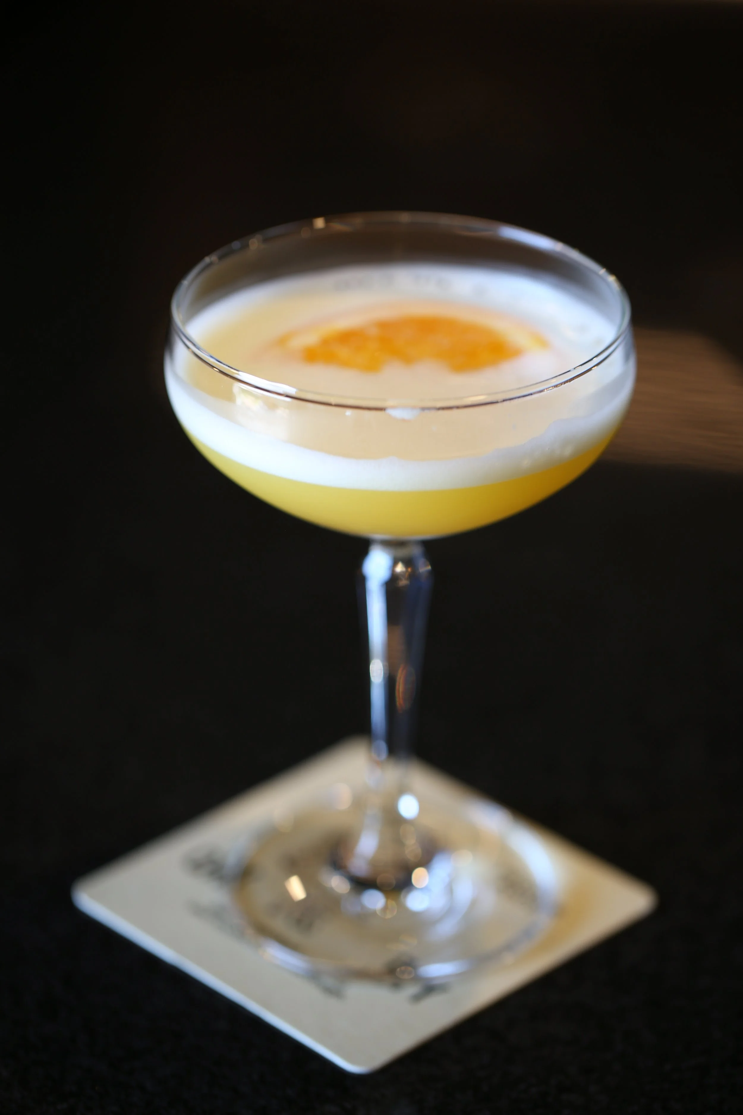 A cocktail in a coupe glass with a yellowish drink, topped with foam and a lemon twist garnish.