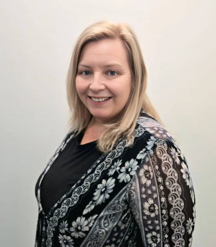 Smiling woman with short reddish hair standing indoors in front of a large window with trees outside, wearing a leopard print top and black pants.
