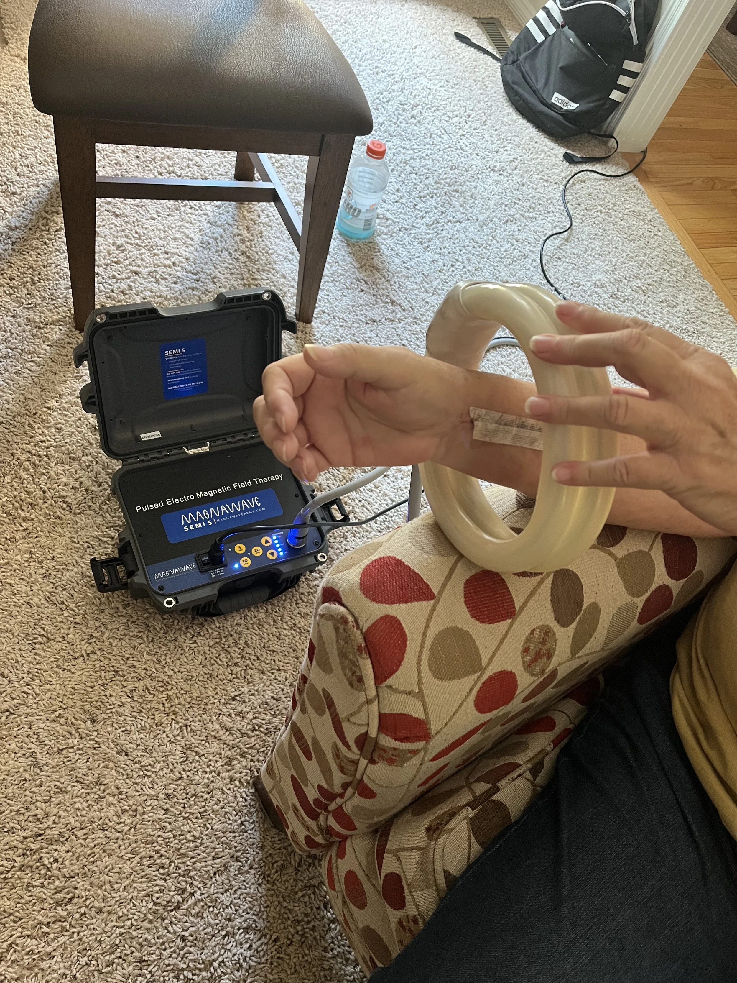 Person sitting on a floral upholstered chair, holding a PEMF treatment device with medical tape on their arm, connected to a portable pulsed electro magnetic field therapy device on the floor.