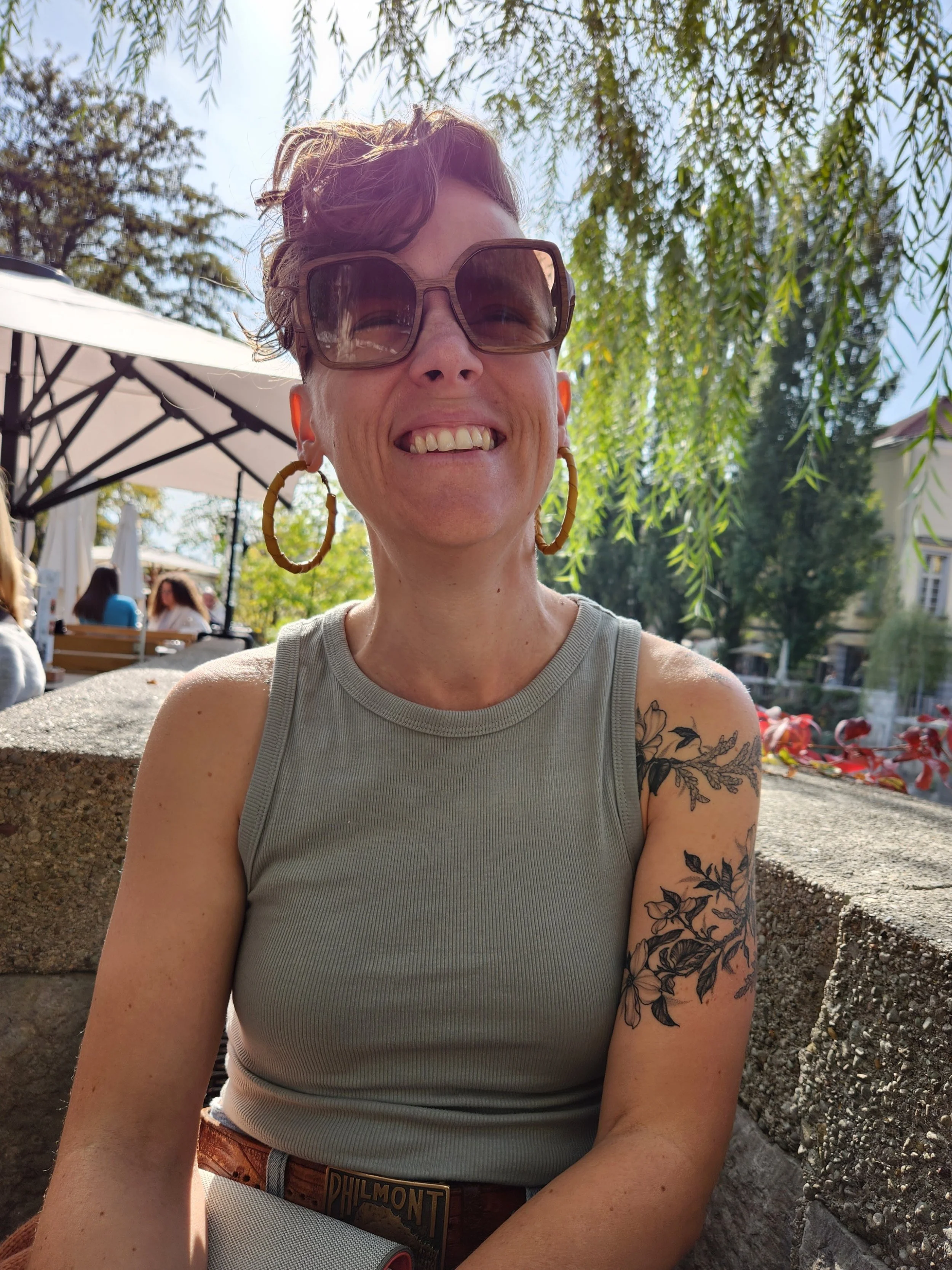 Megan Rossi, Art and Alchemy co-facilitator at The Unlearning Studio, smiling outdoors at a café table wearing sunglasses beneath an umbrella