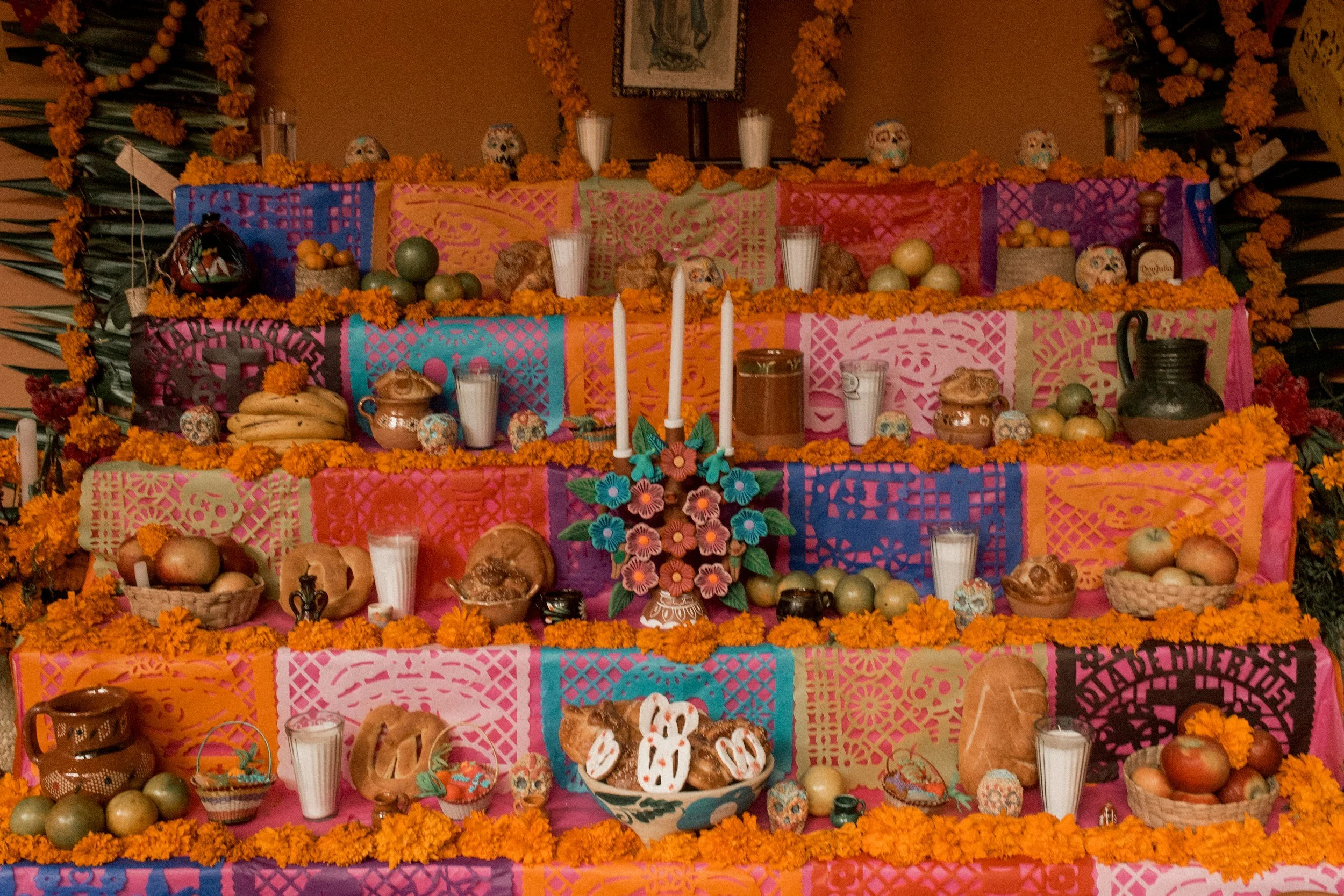 Day of the Dead altar decorated with marigold flowers, traditional artwork, skull figurines, candles, and offerings such as fruits and baked goods, against colorful papel picado banners.
