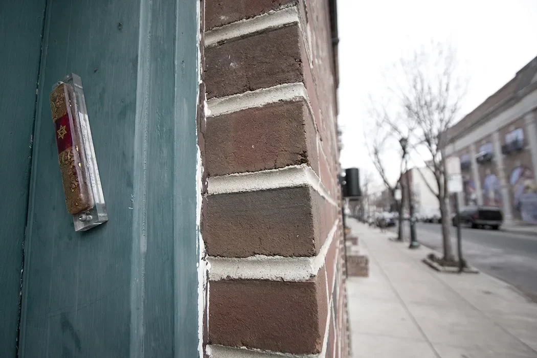 Close-up of a brick wall with mortar lines, adjacent to a green wooden door with a small package attached to it, along a city sidewalk with bare trees and buildings in the background.