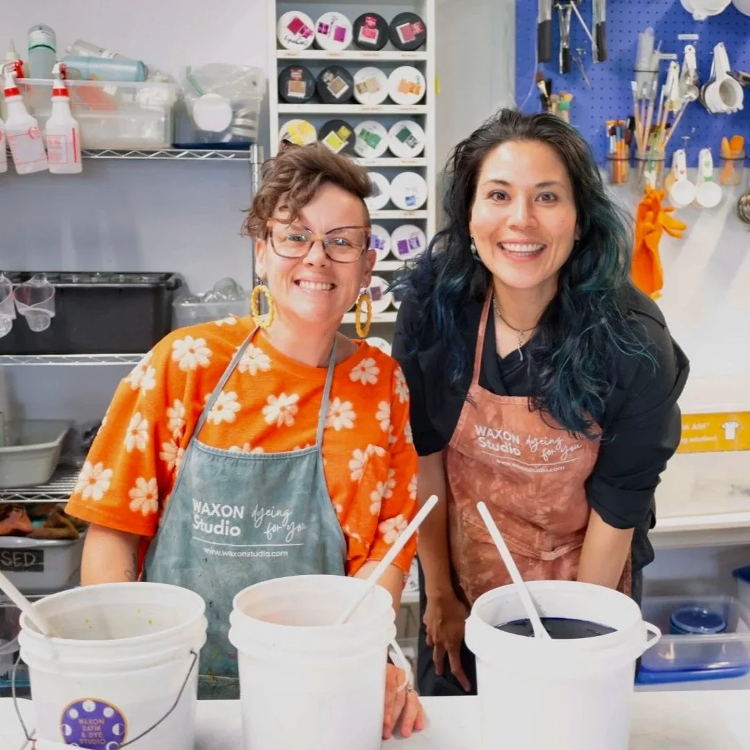Two women smiling in a craft or art studio, one wearing glasses and an orange floral shirt, the other with dyed blue hair and wearing a black shirt, in front of buckets of black and white paint on a work table.
