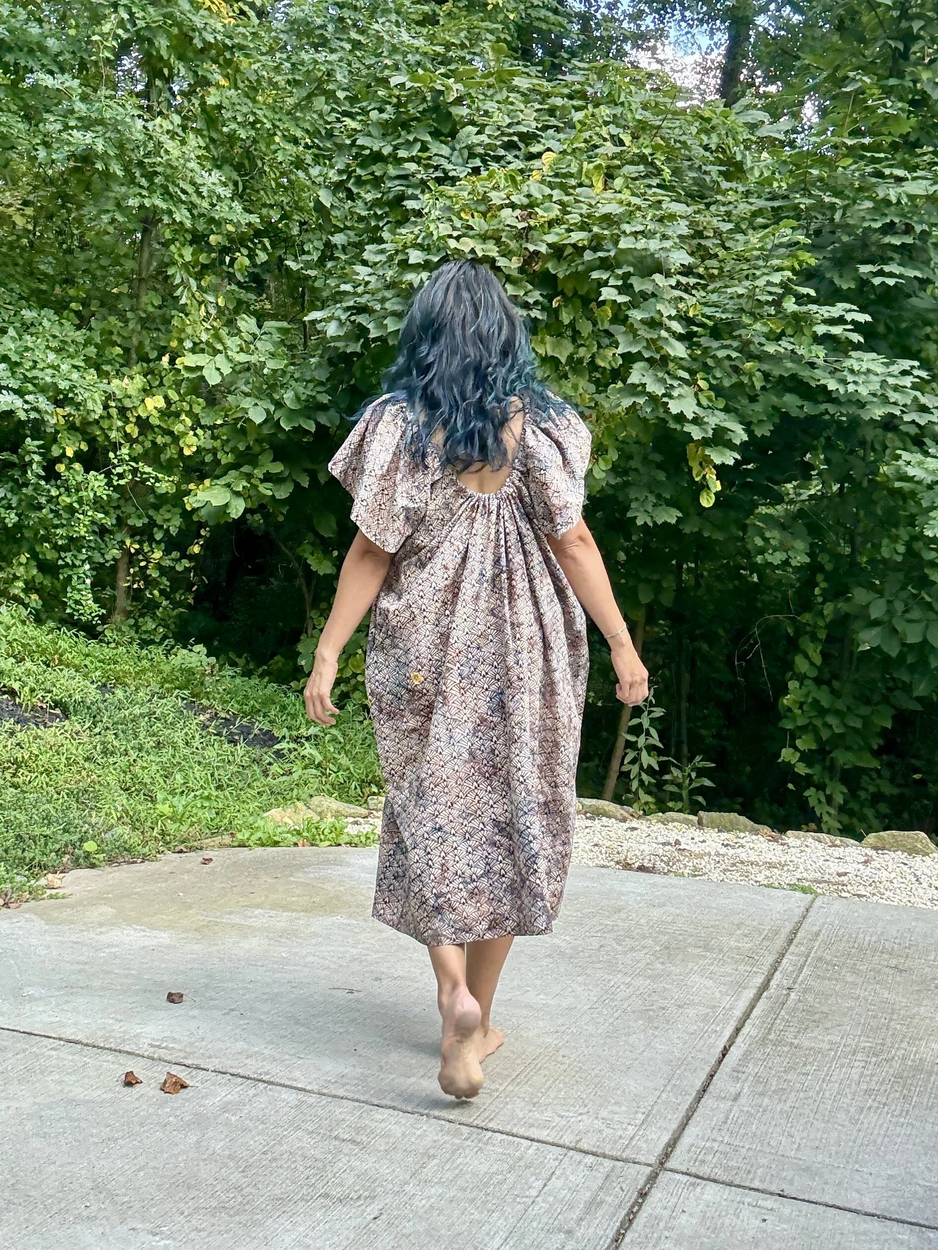 Alice Chen in a summer dress walking away from the camera toward the woods, an image of quiet courage, forward movement and following what calls you
