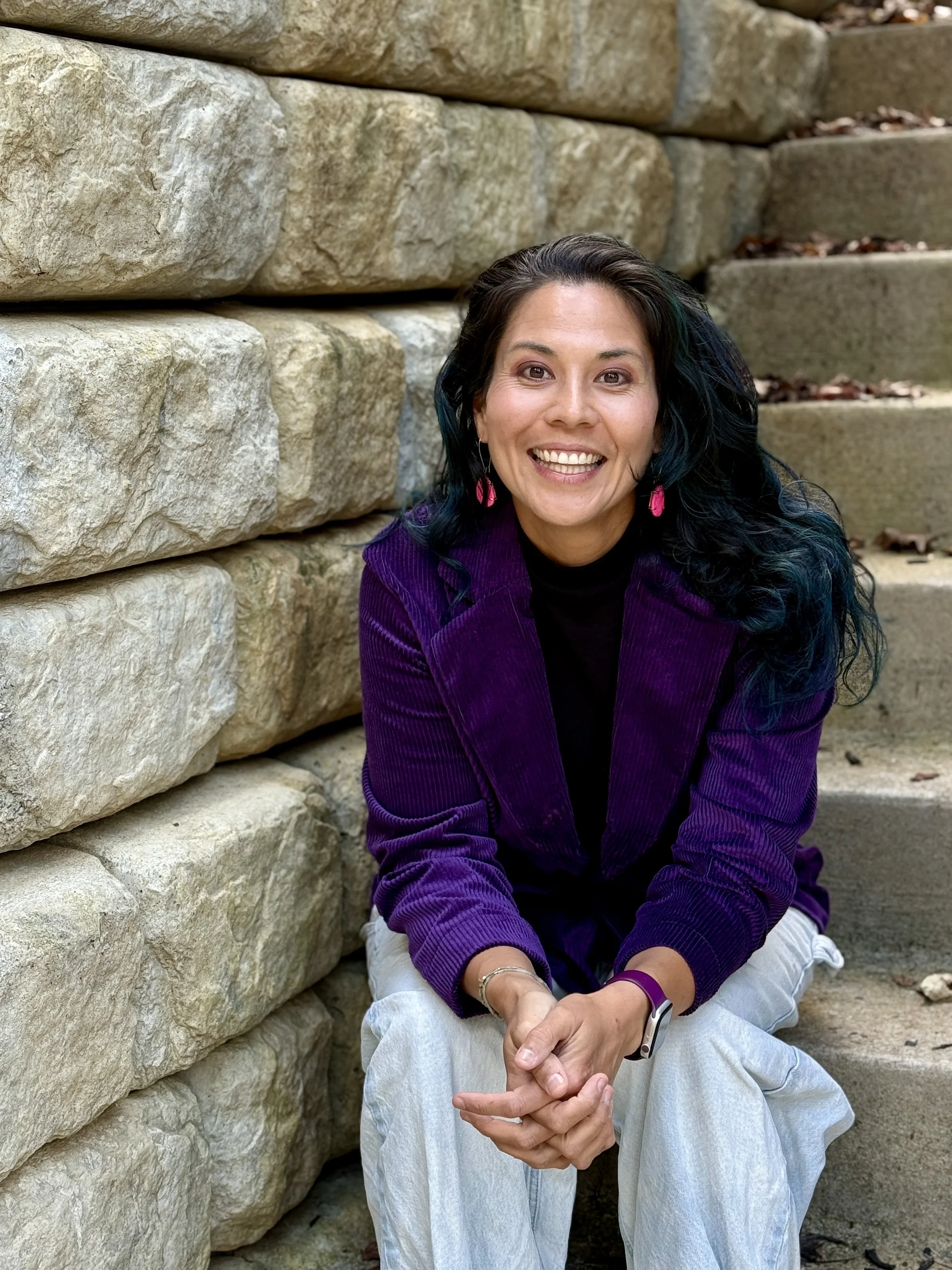 Alice Chen, founder of The Unlearning Studio, smiling on outdoor stairs wearing a purple velvet jacket and neon pink earrings — bold, warm and unmistakably herself