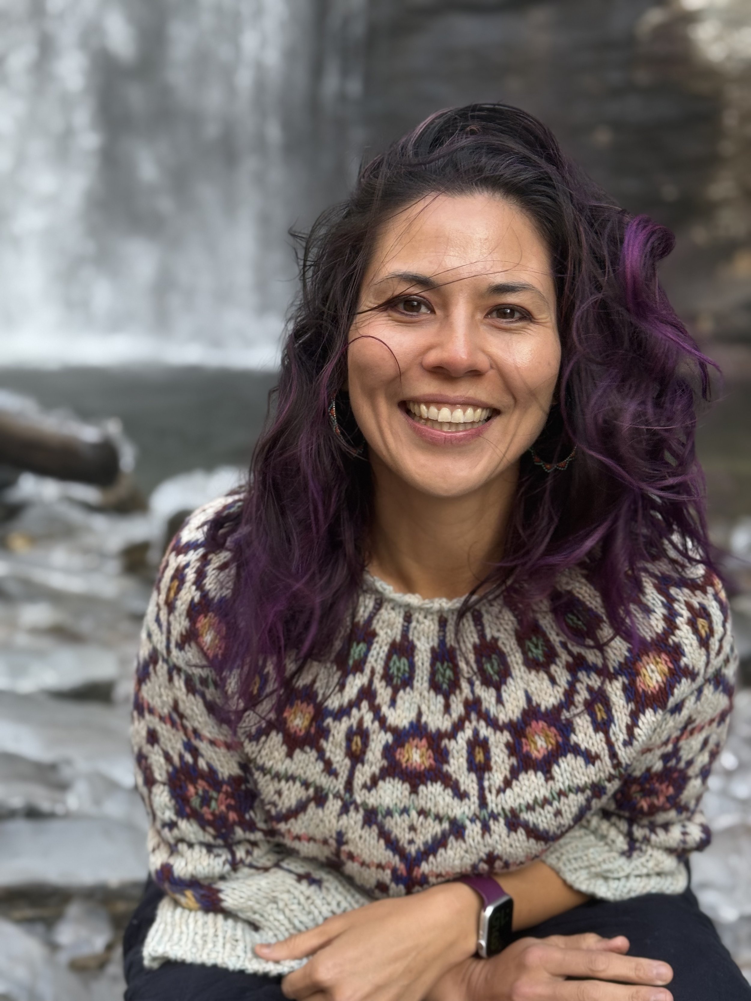 Alice Chen smiling in a handknit sweater with a waterfall cascading behind her, embodying the creative joy and natural aliveness at the heart of her work