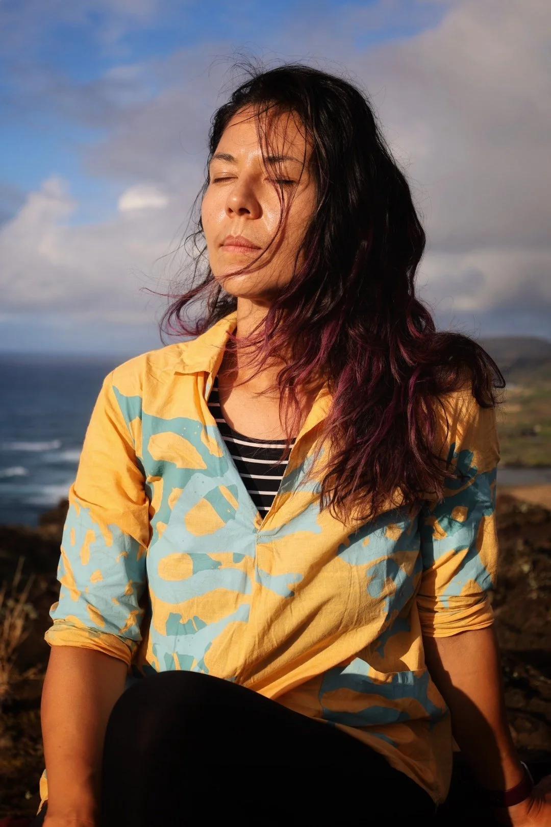 Alice Chen in an orange and teal Hawaiian shirt with eyes closed, face turned toward the sunrise — a moment of stillness, presence and reclamation