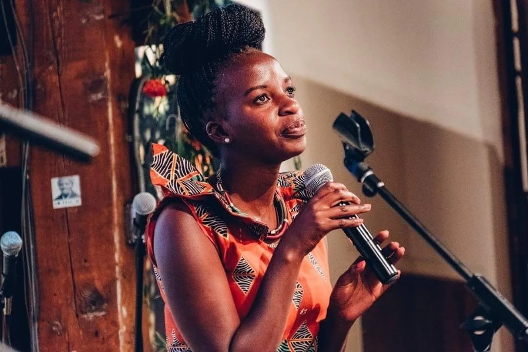A woman holding a microphone and speaking or singing indoors, with a wooden wall and plants in the background.