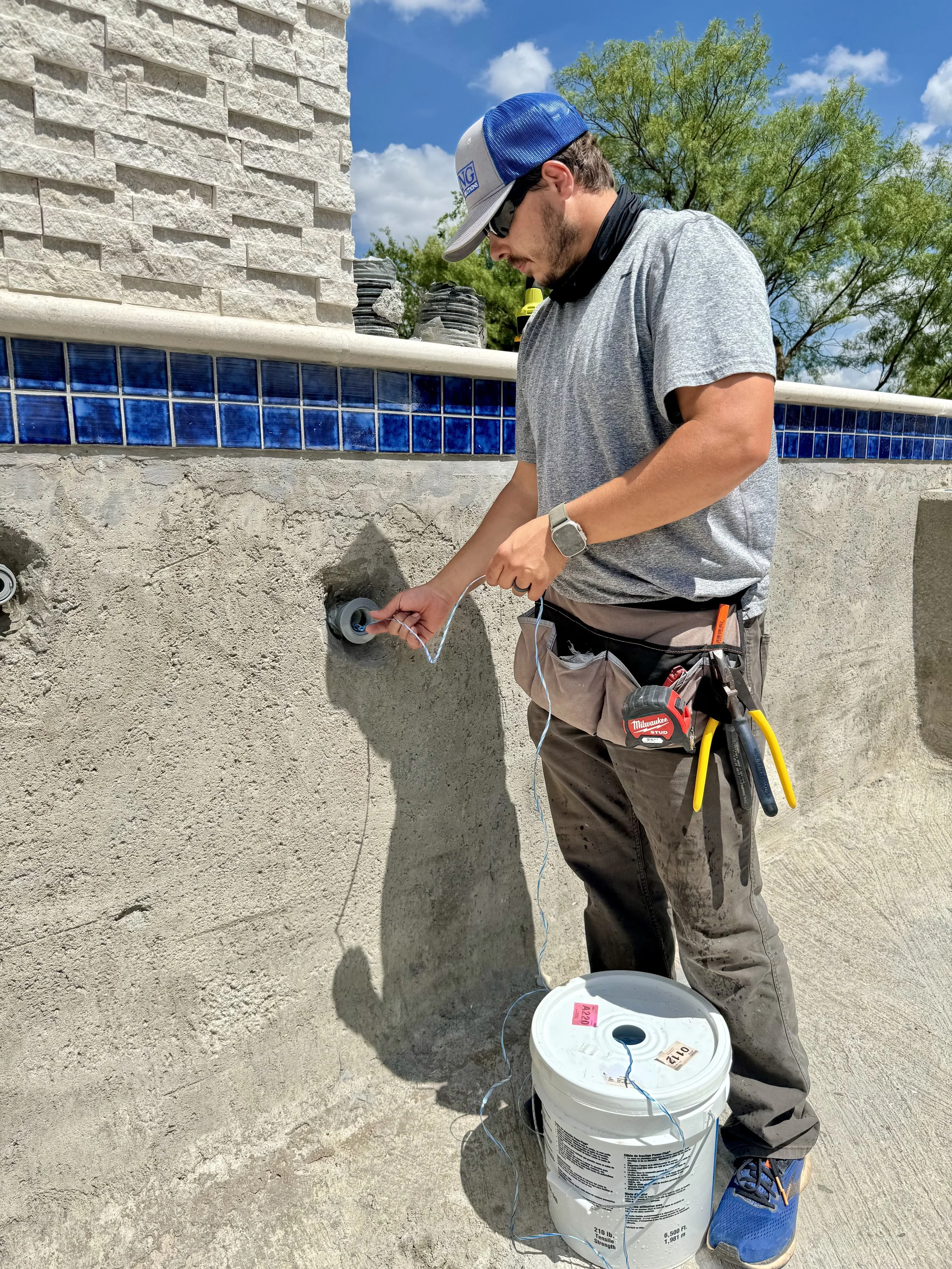 A man working outdoors installing or repairing a wall fixture with wires, standing next to a bucket and wearing casual work clothes, a cap, and safety tools.