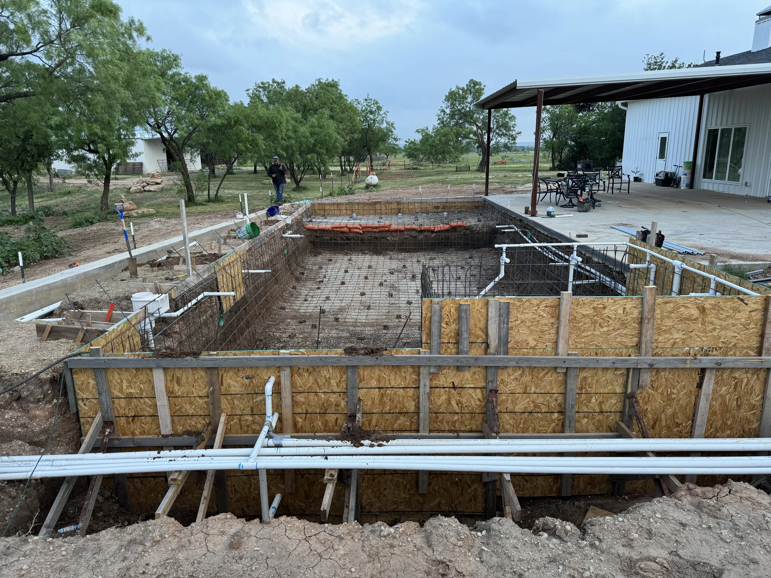 Construction site with a dug-out area with rebar and plumbing, surrounded by wooden and metal forms, outside a white building with furniture on a patio, and green trees in the background.