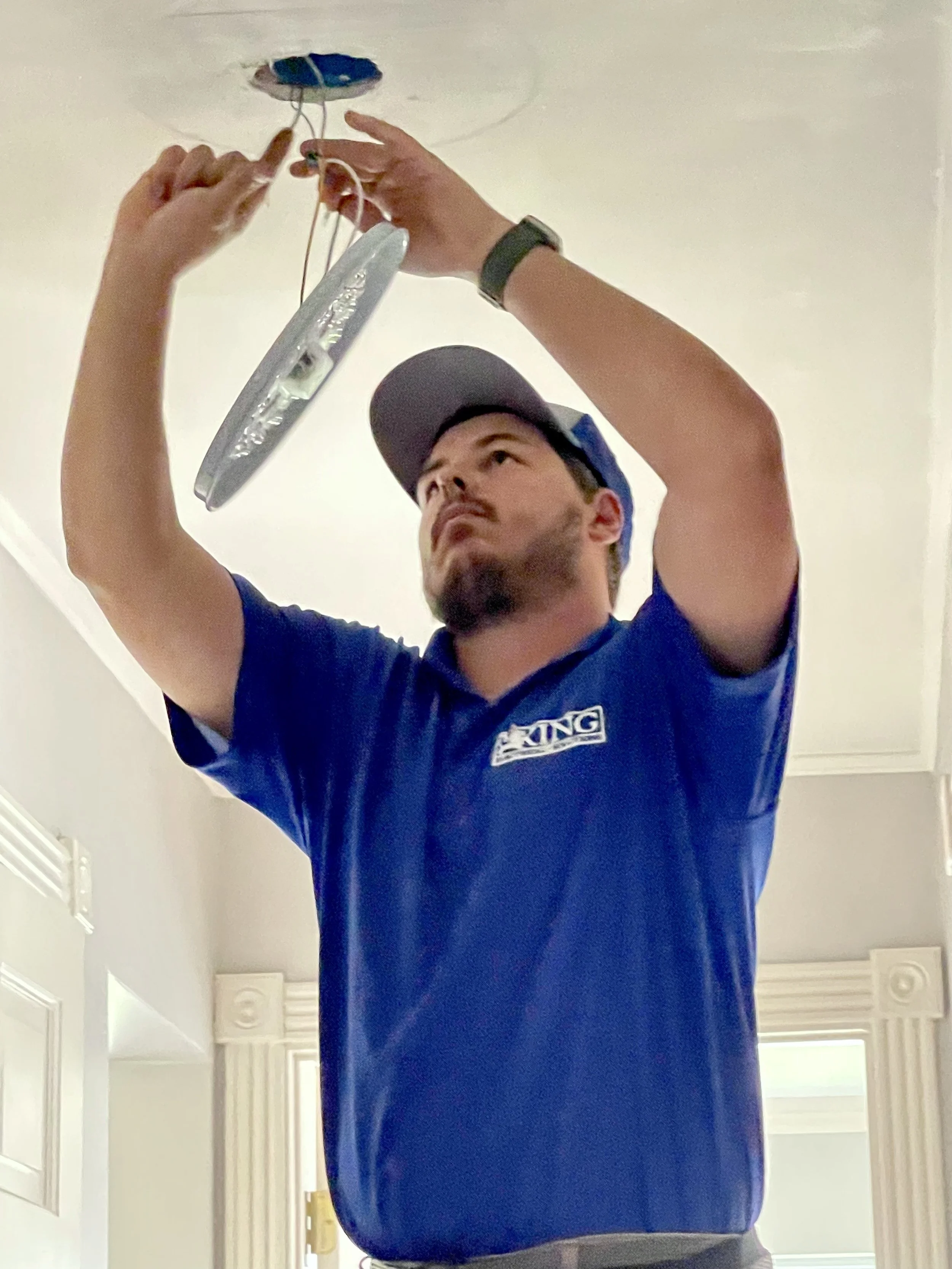 A man installing a ceiling light fixture, wearing a blue uniform and a cap, is reaching up and working on electrical wires near an opening in the ceiling inside a house.