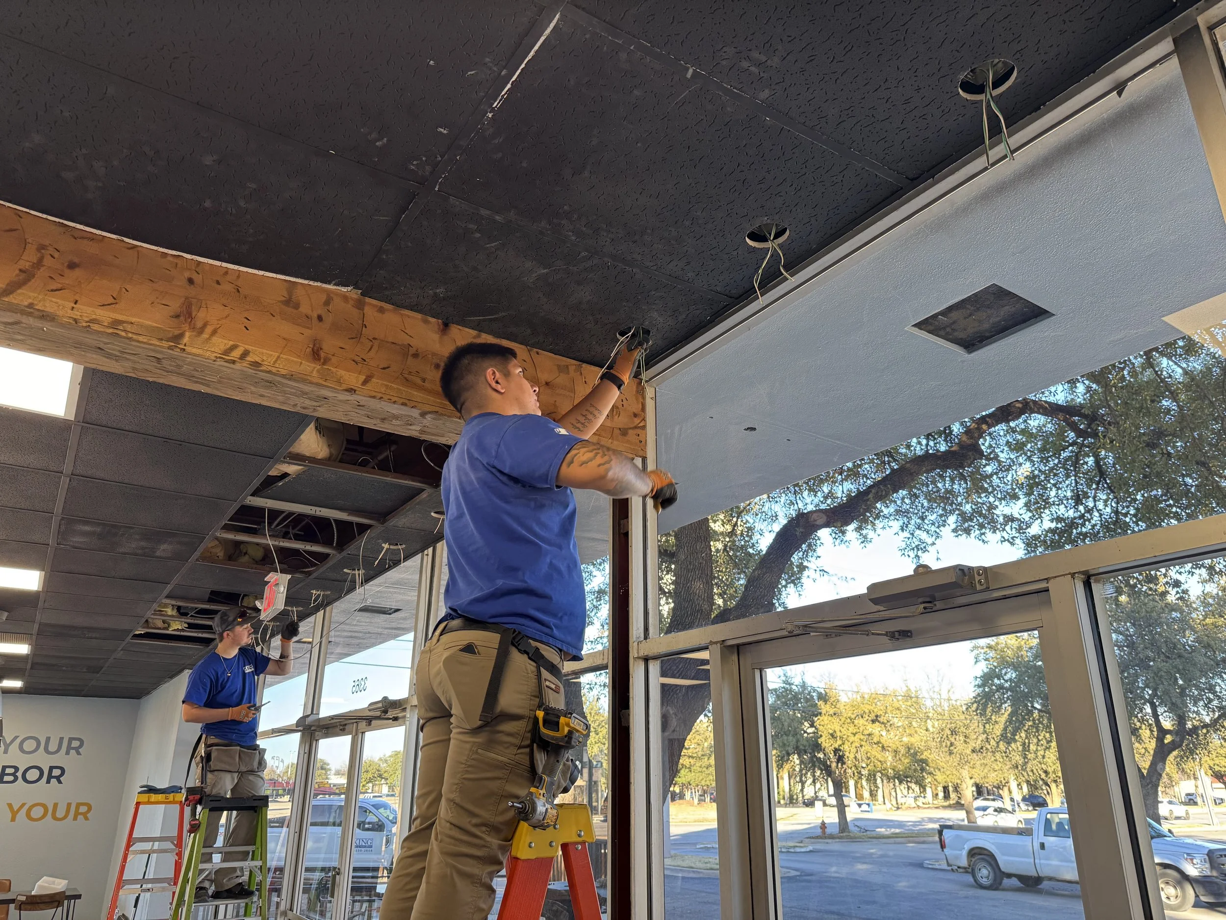 Two workers installing ceiling panels in a commercial space. One is on a ladder, the other is standing on a stepladder, both working on the ceiling installation near large windows with trees outside.