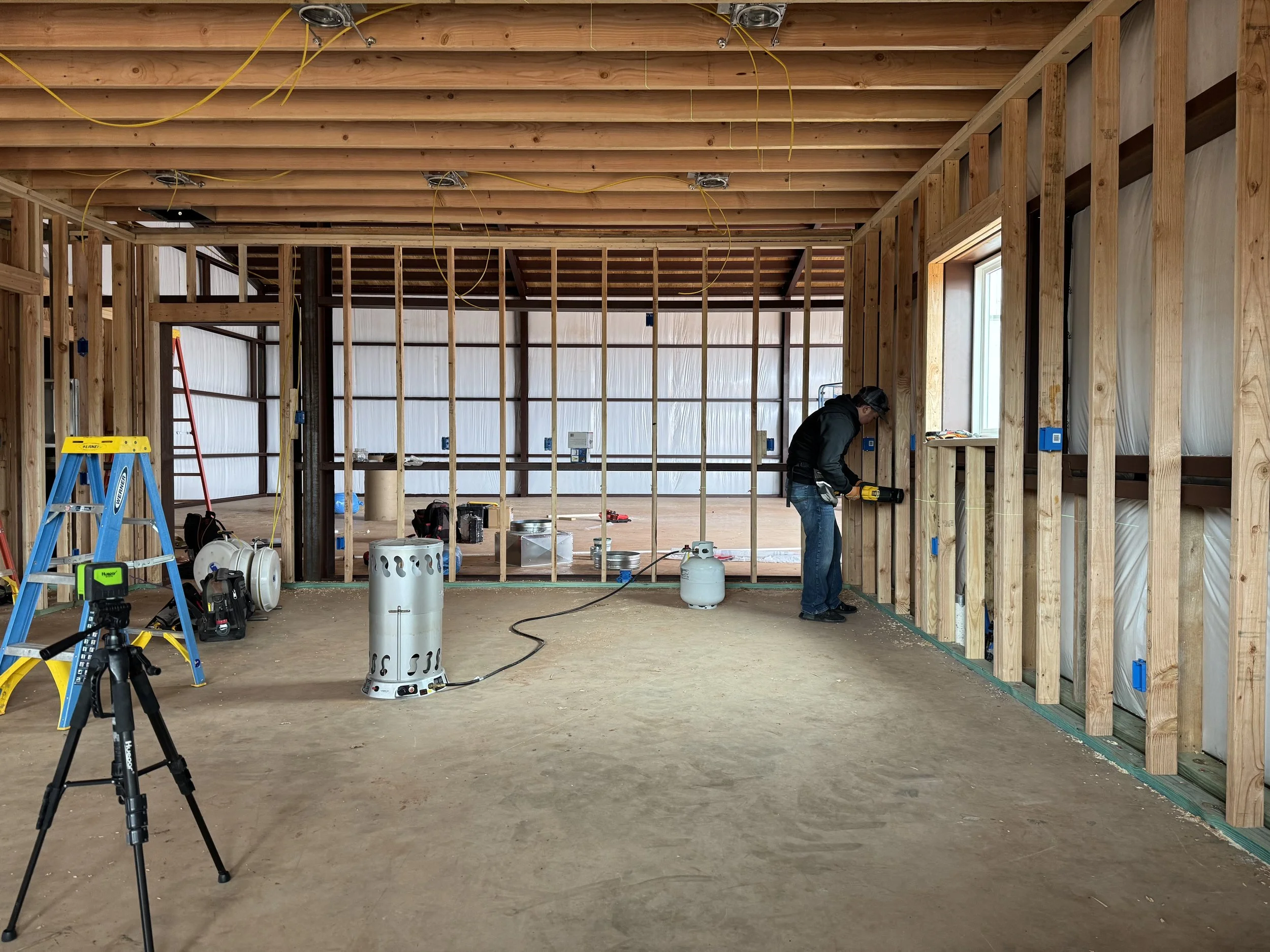 Interior of a house under construction, showing wooden framing, construction tools, and a worker using a power drill.