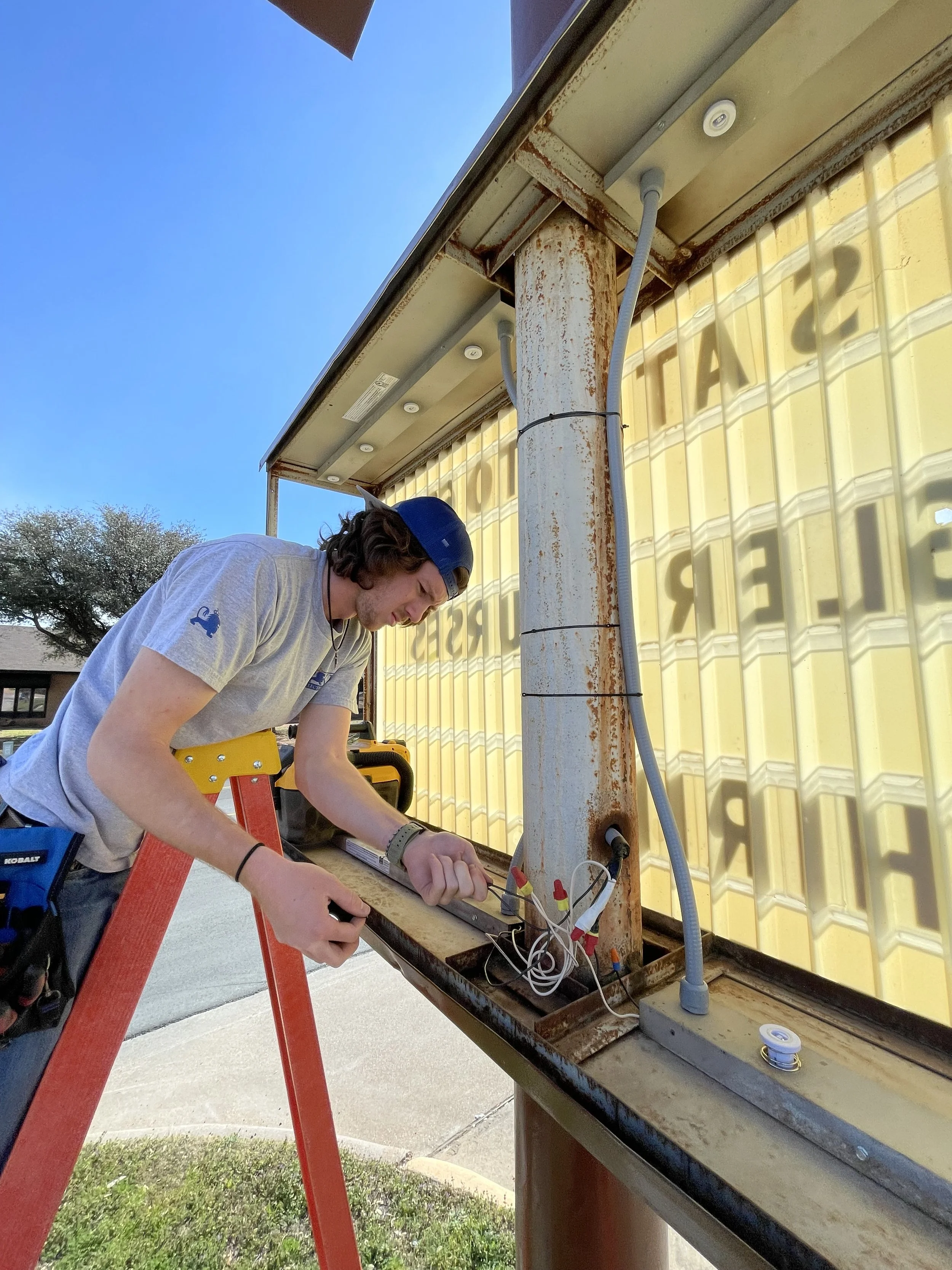 Young man working on electrical wiring at an outdoor site with a rusty metal post and yellow insulated panel in the background.