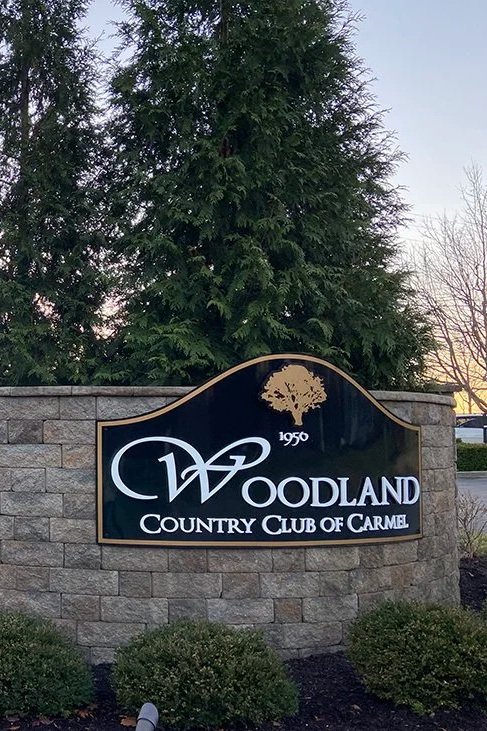 Sign for Woodland Country Club of Carmel, established in 1950, with a large tree graphic and surrounded by greenery.
