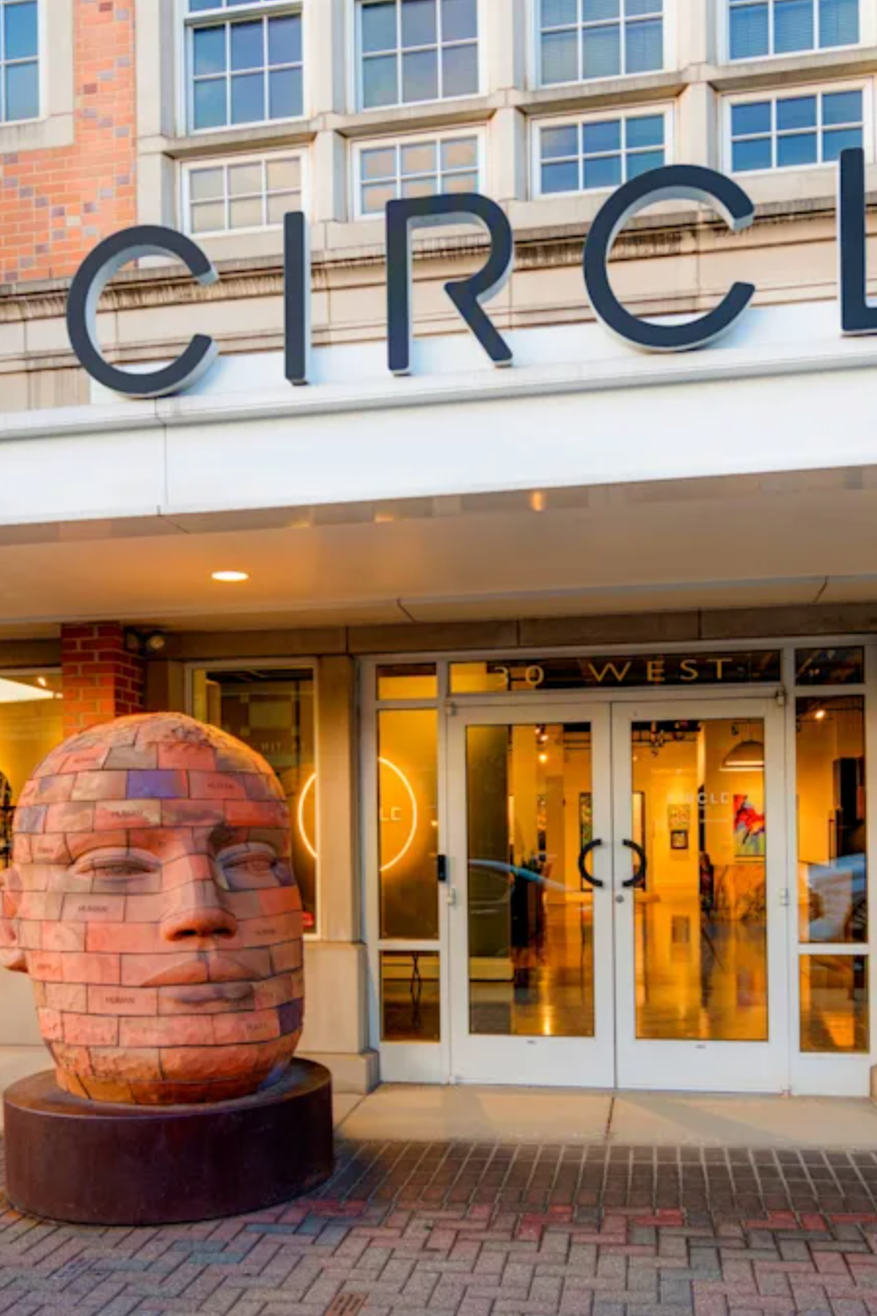 Entrance of a building with large glass doors, a sculpture of a human head made of bricks outside, and the partial word 'CIRC' displayed above.