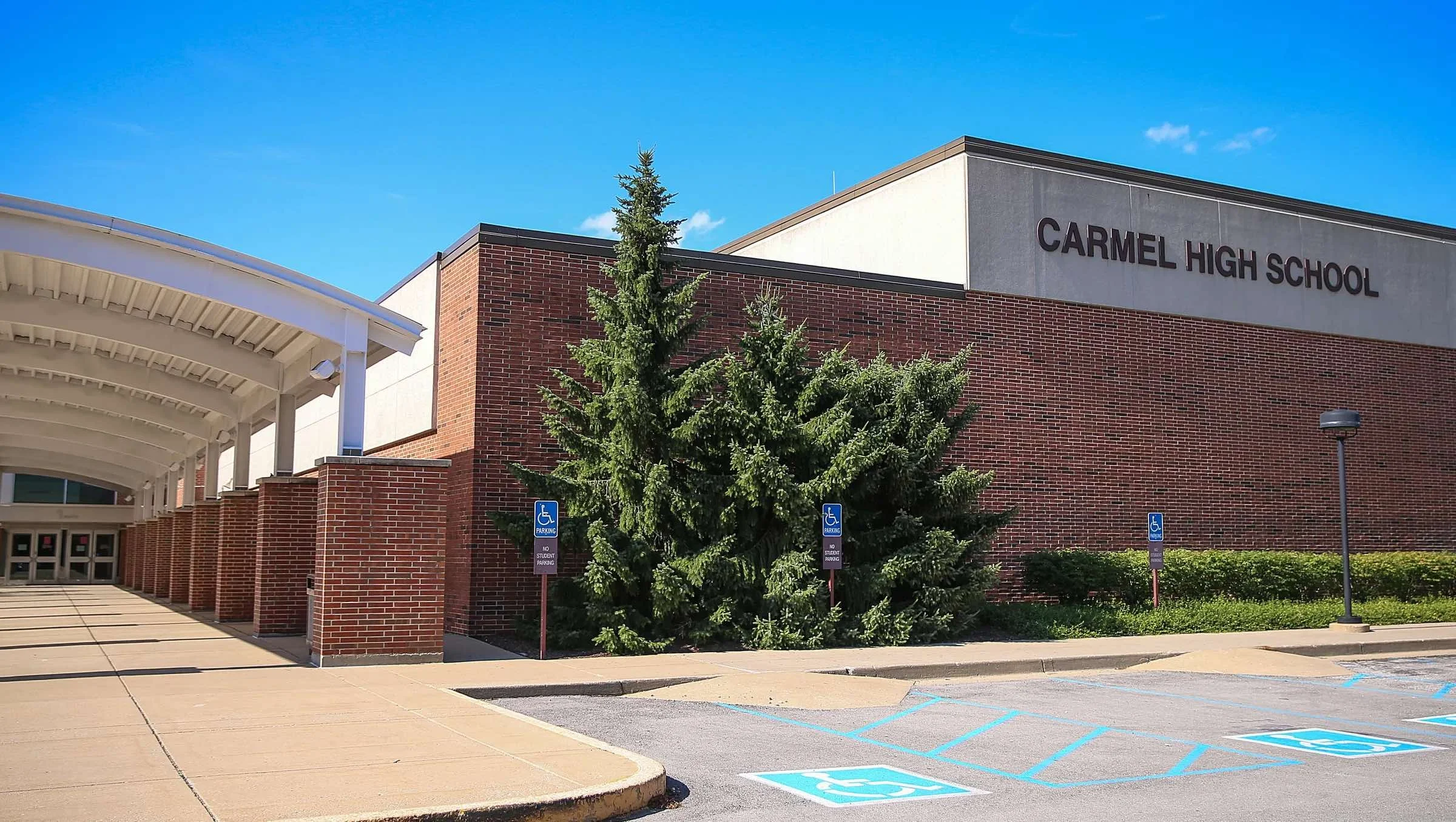 Exterior view of Carmel High School building with red brick walls, a concrete section with the school's name, parking spaces marked for handicapped parking, and a row of green bushes and trees under a blue sky.