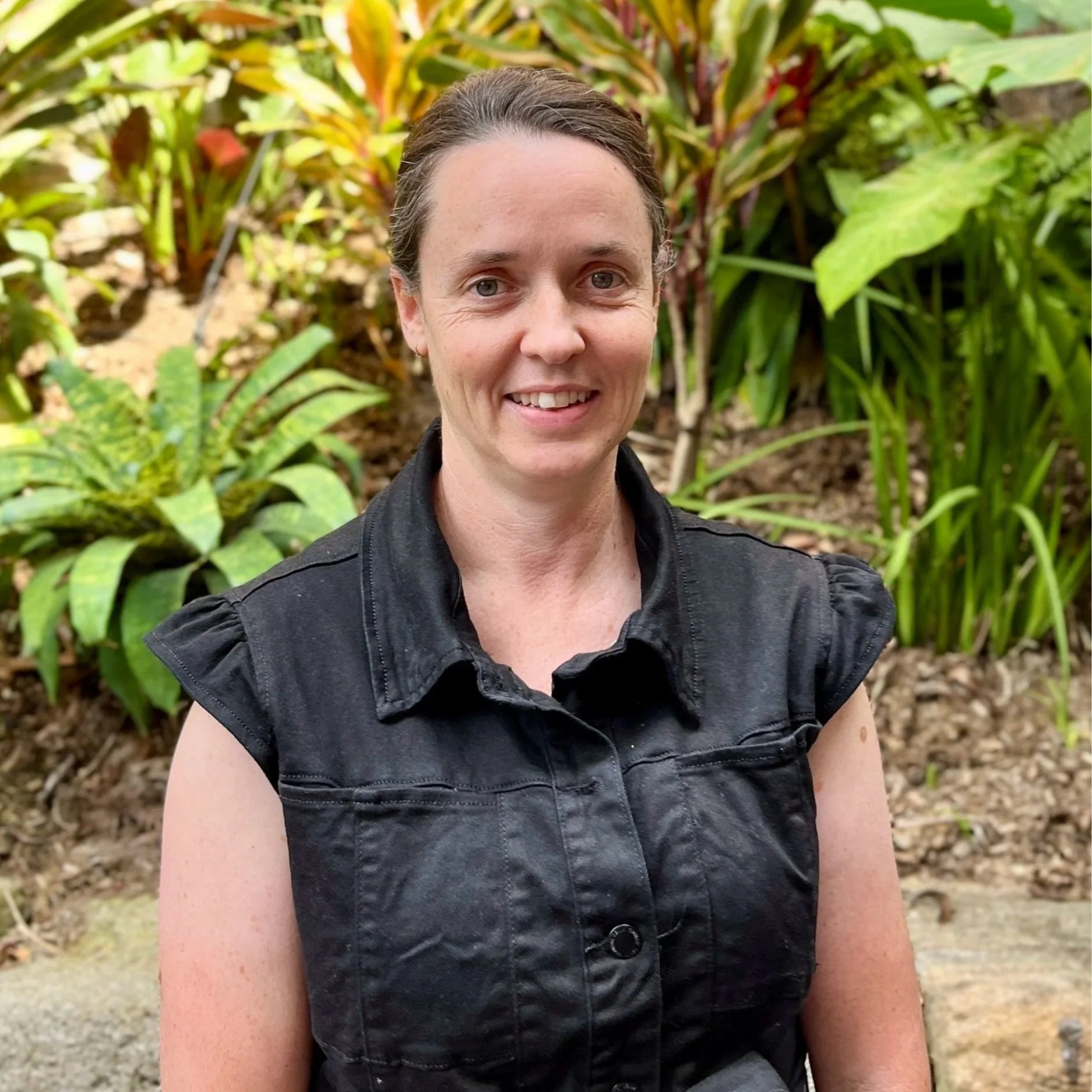 A woman with dark hair, fair skin, and freckles, smiling, wearing a sleeveless black floral top, standing against a plain off-white wall.