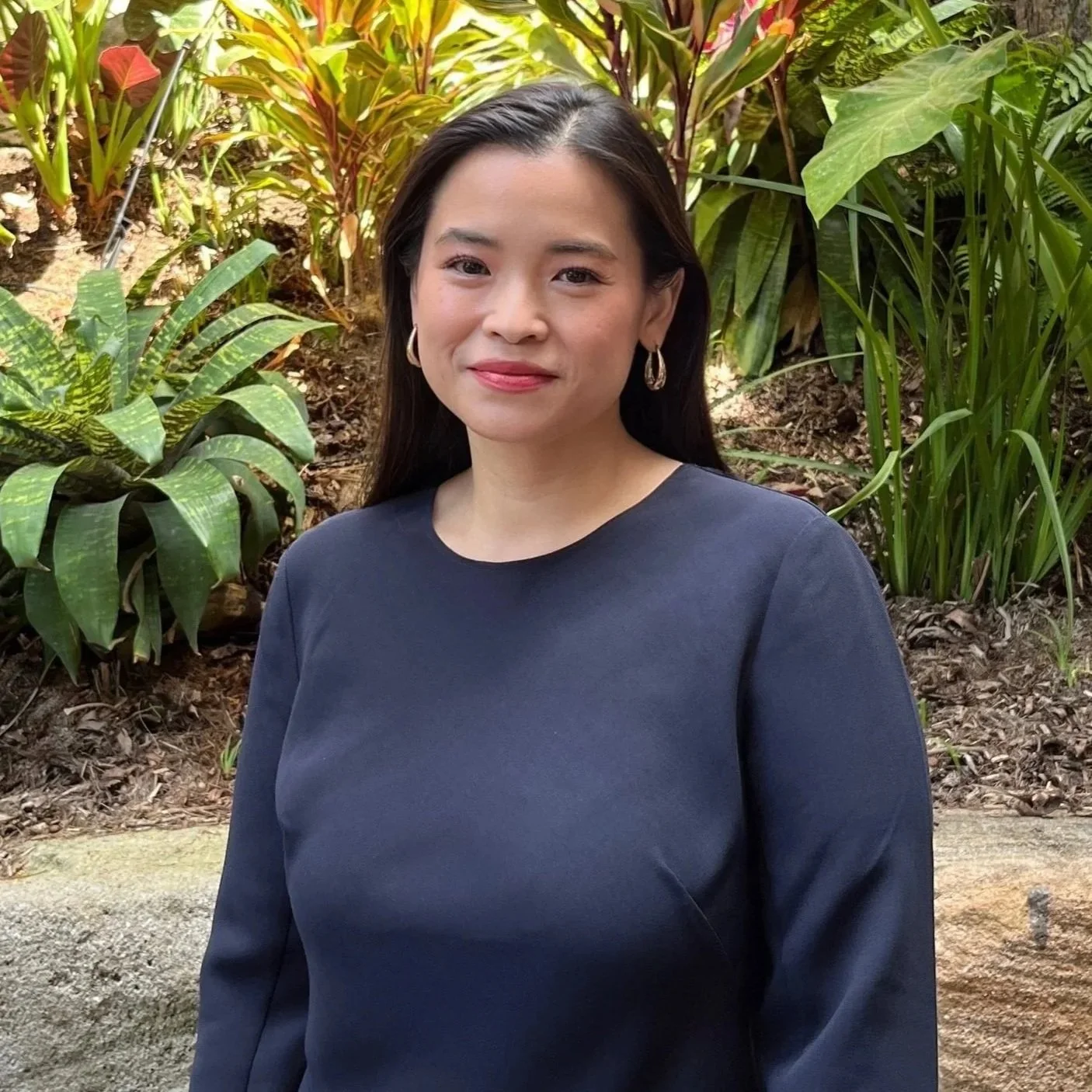 A woman with shoulder-length black hair wearing a dark satin dress and gold jewelry, standing against a plain white background.